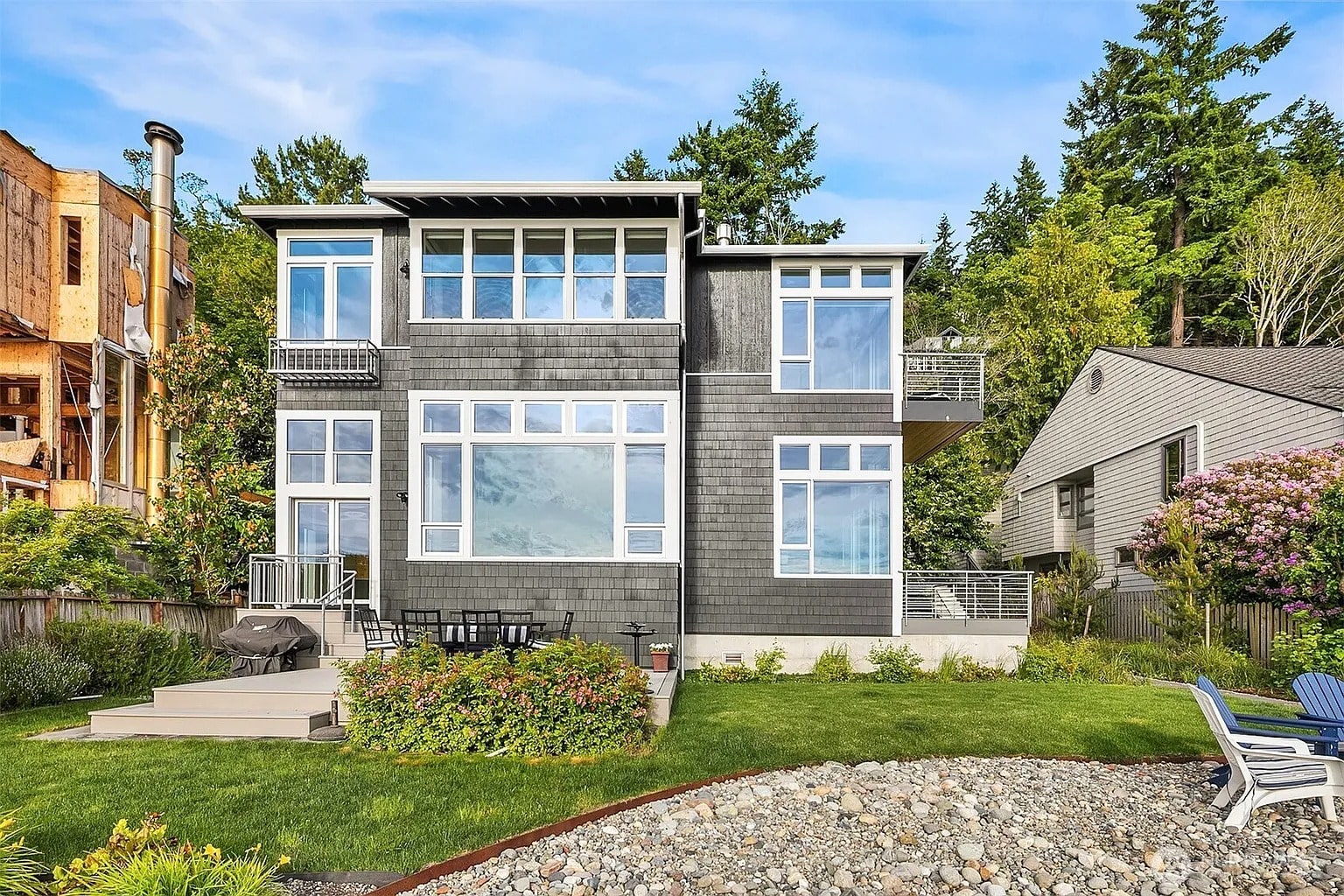 Modern two-story house with large windows, surrounded by greenery and neighboring homes.