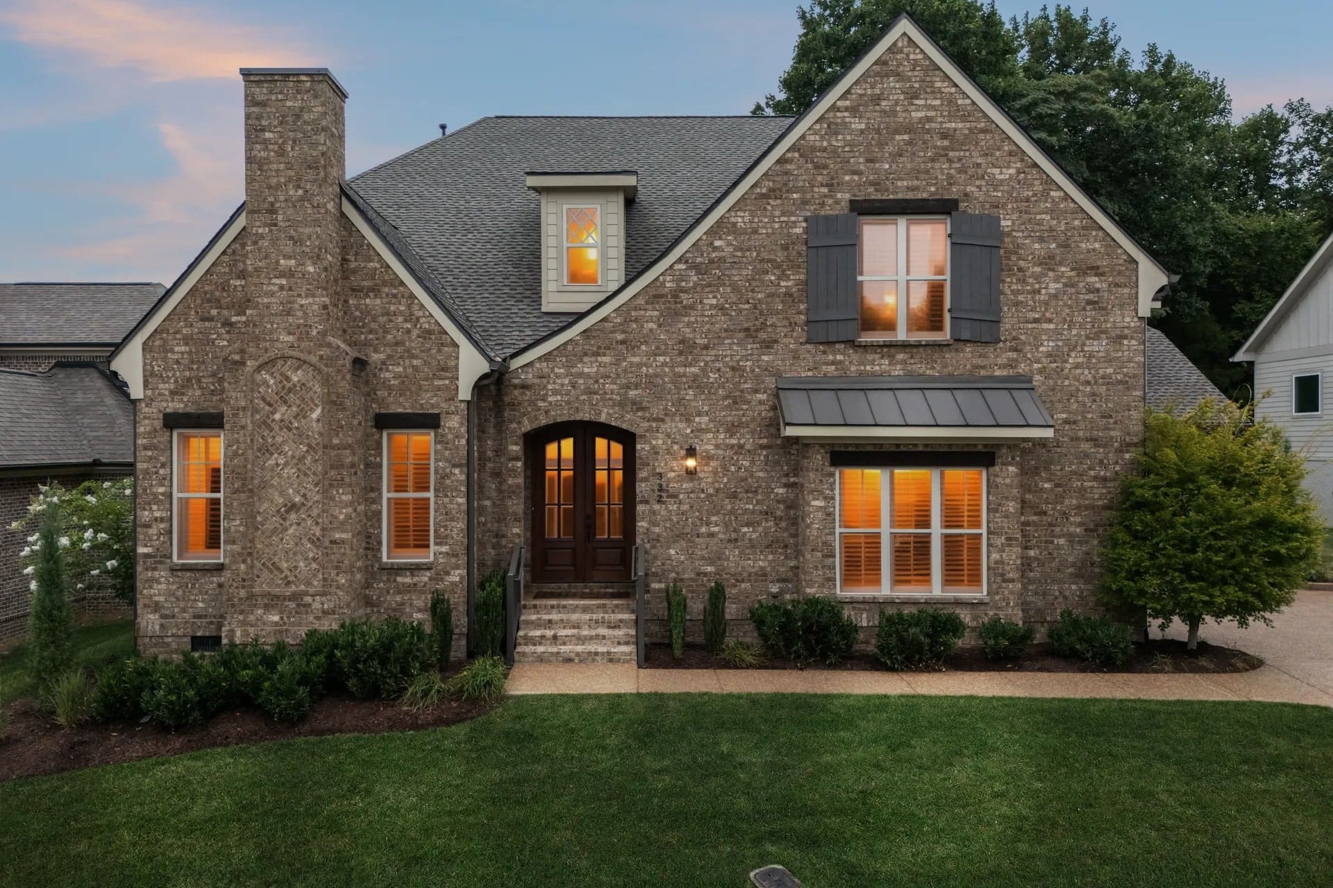 Brick house with arched doorway, lit windows, and manicured lawn at dusk.