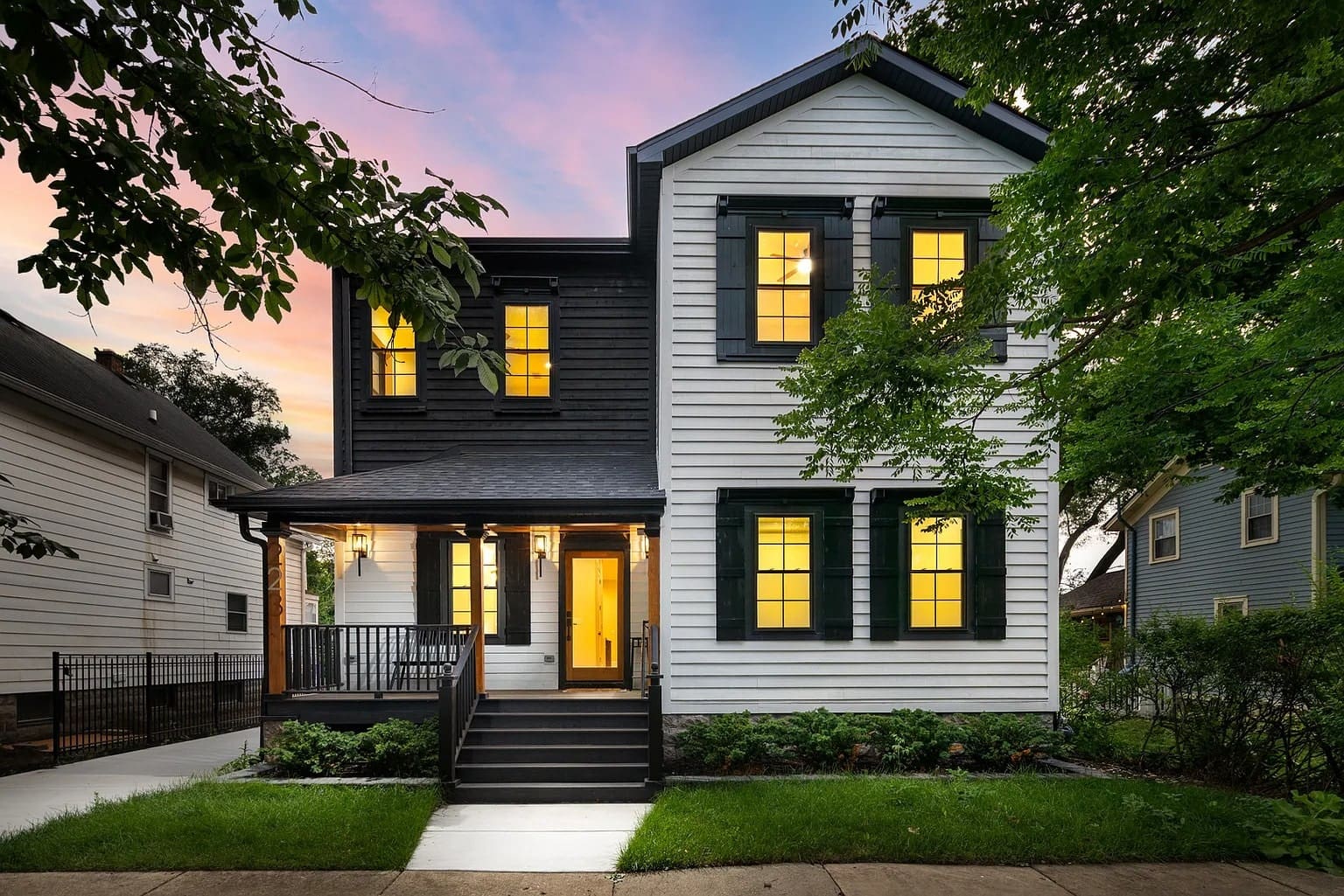 Two-story house with black and white exterior, illuminated windows, and surrounding trees at sunset.