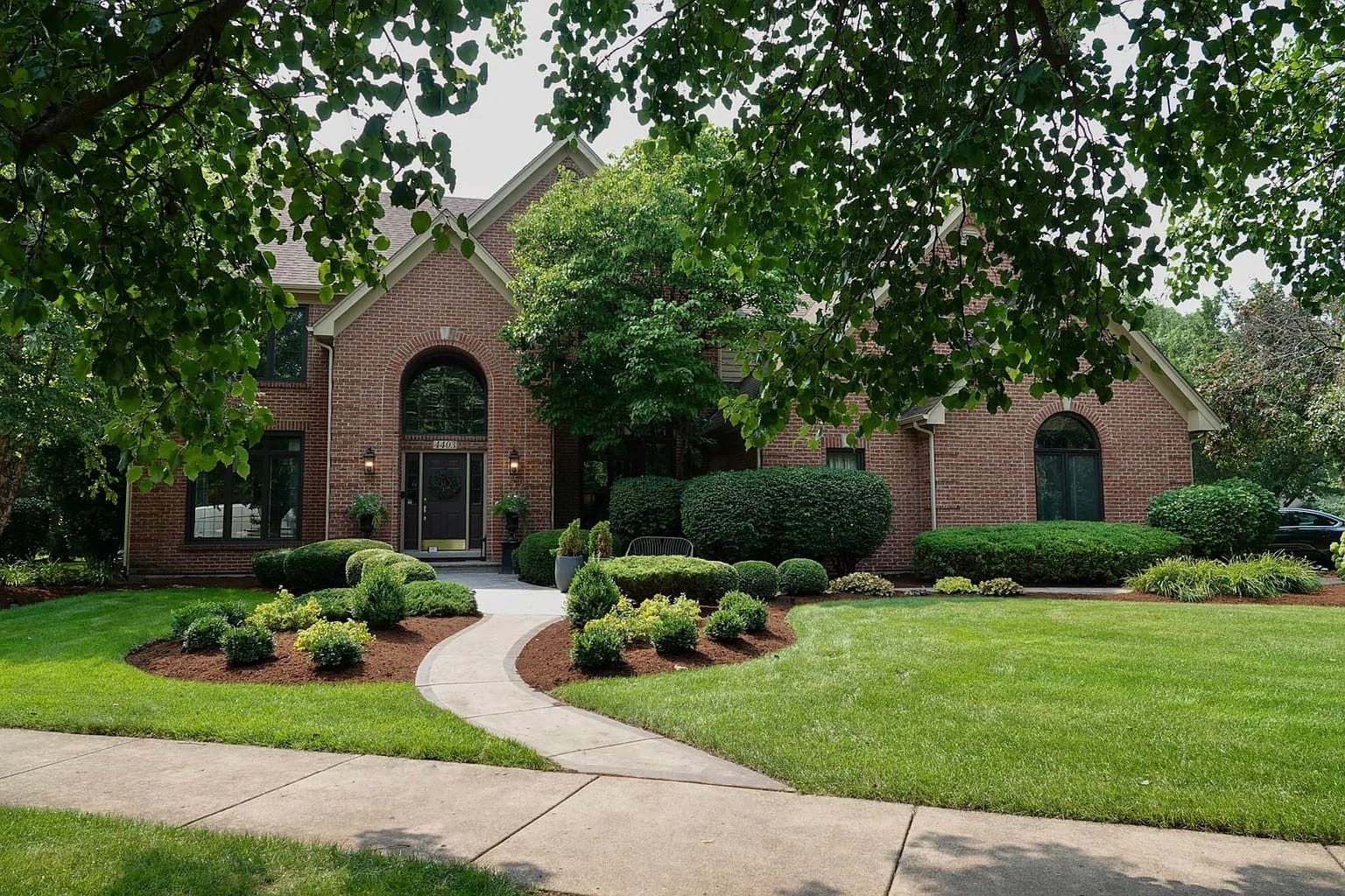 Brick house with arched windows, surrounded by manicured lawn and shrubs, under a tree canopy.