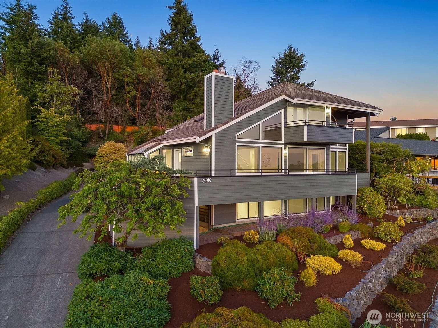 Modern two-story house with large windows, surrounded by trees and landscaped garden.