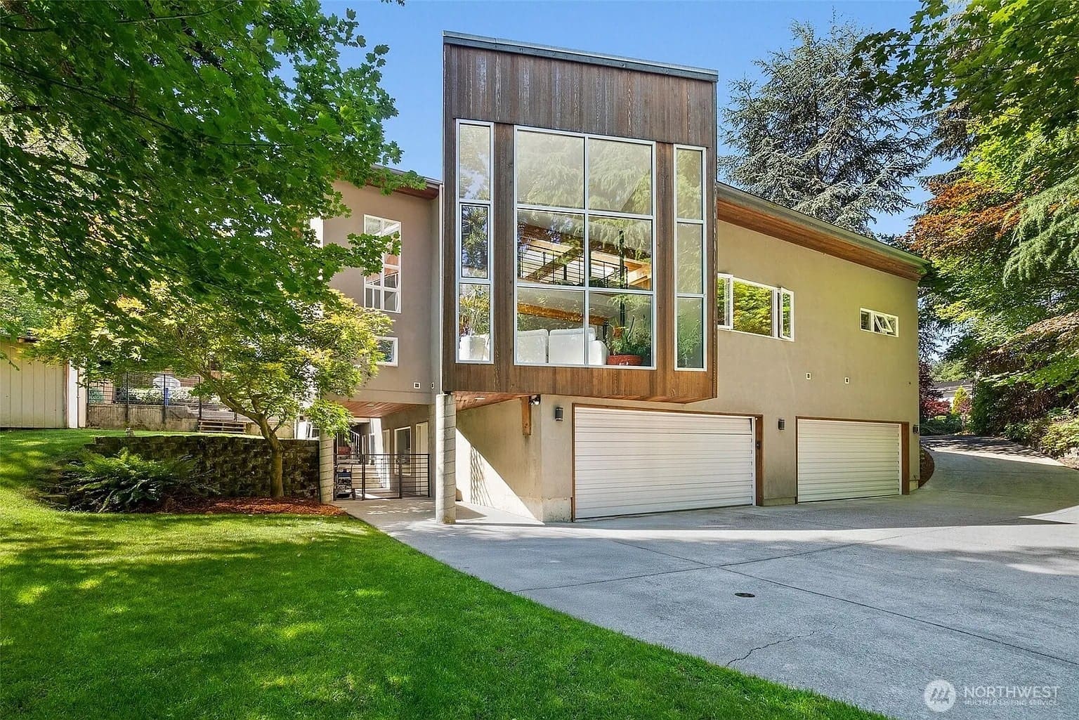Modern house with large glass windows, three-car garage, surrounded by trees and greenery.