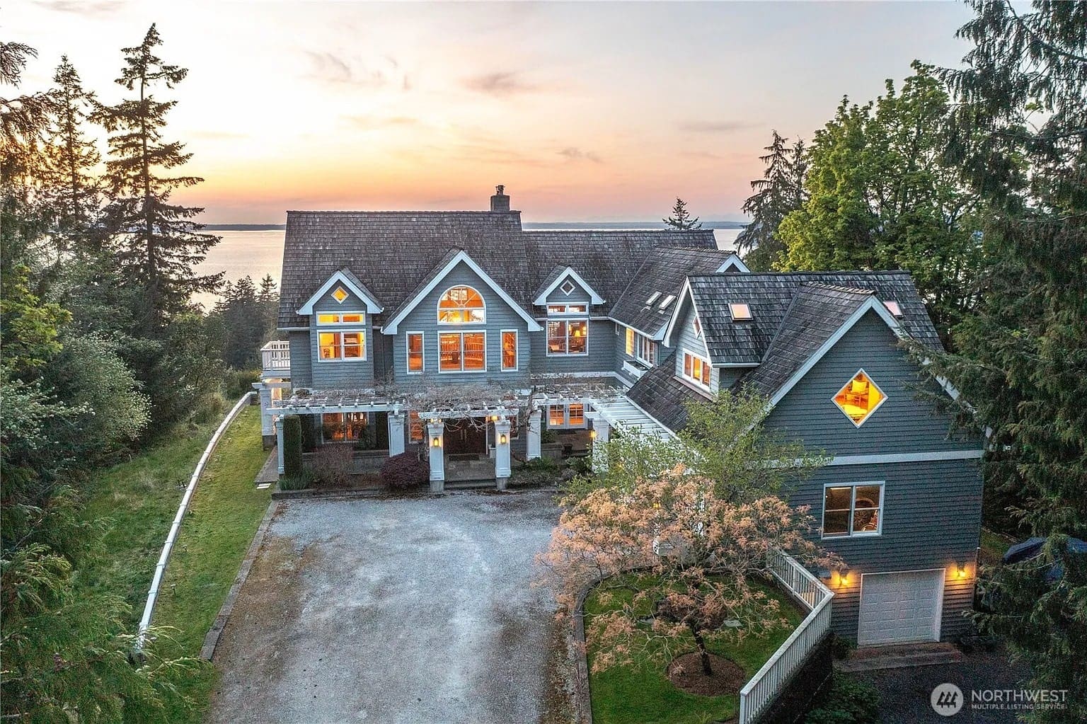 Large coastal house with multiple gables, surrounded by trees, overlooking a sunset view of the water.