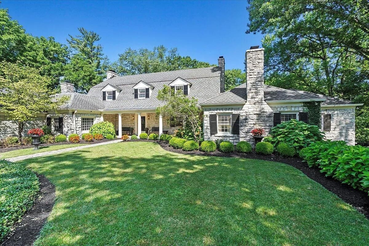 Large stone house with a well-maintained lawn, shrubs, and trees under a clear blue sky.