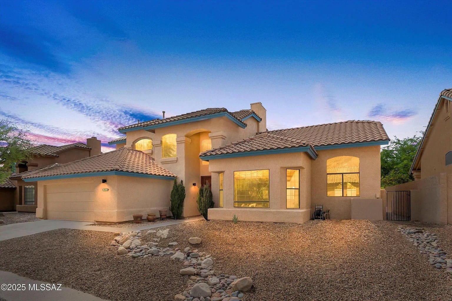 Two-story stucco house with tile roof, illuminated at dusk, surrounded by a gravel yard.