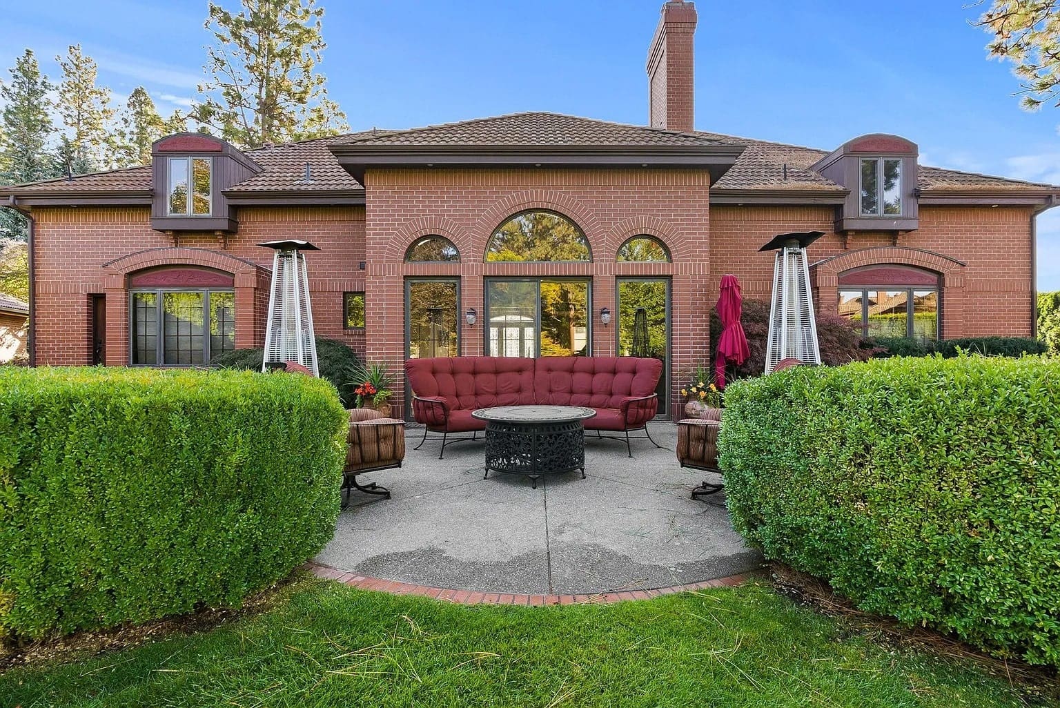 Brick house with large windows, patio furniture, and outdoor heaters, surrounded by greenery.