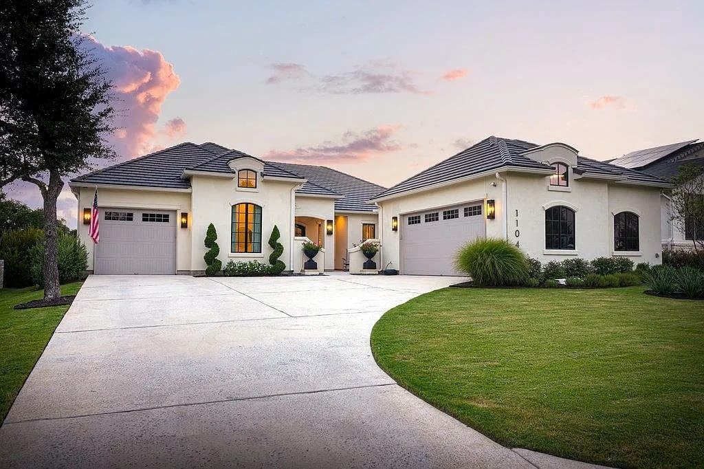 Modern single-story house with a large driveway, manicured lawn, and American flag at sunset.