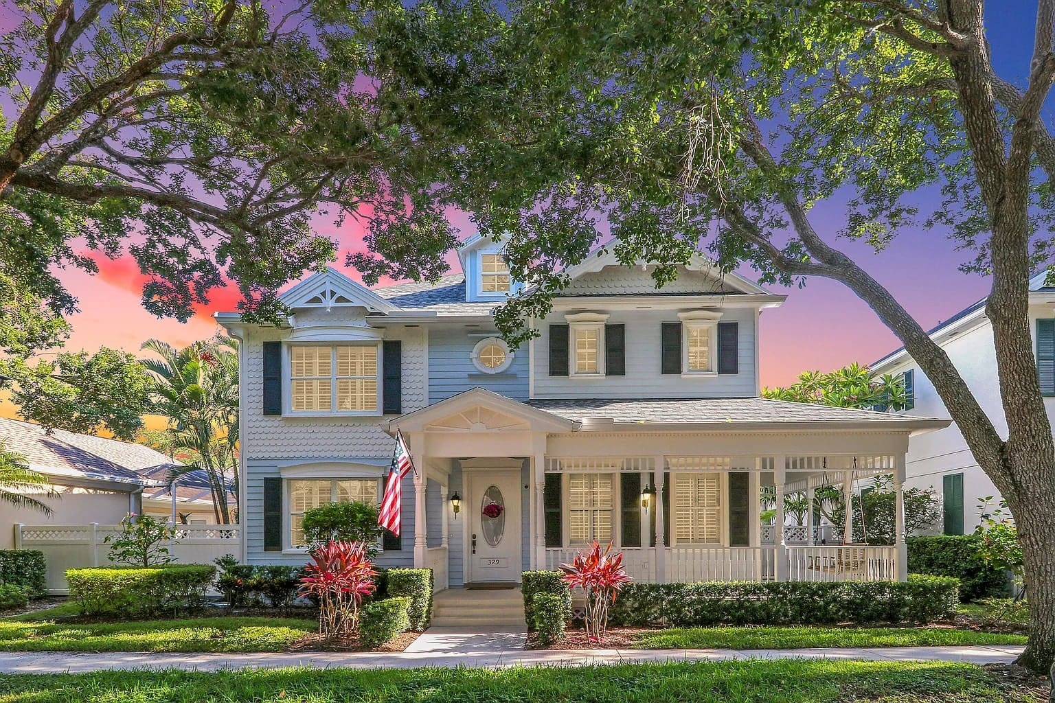 Two-story house with porch, American flag, surrounded by trees, under a colorful sunset sky.