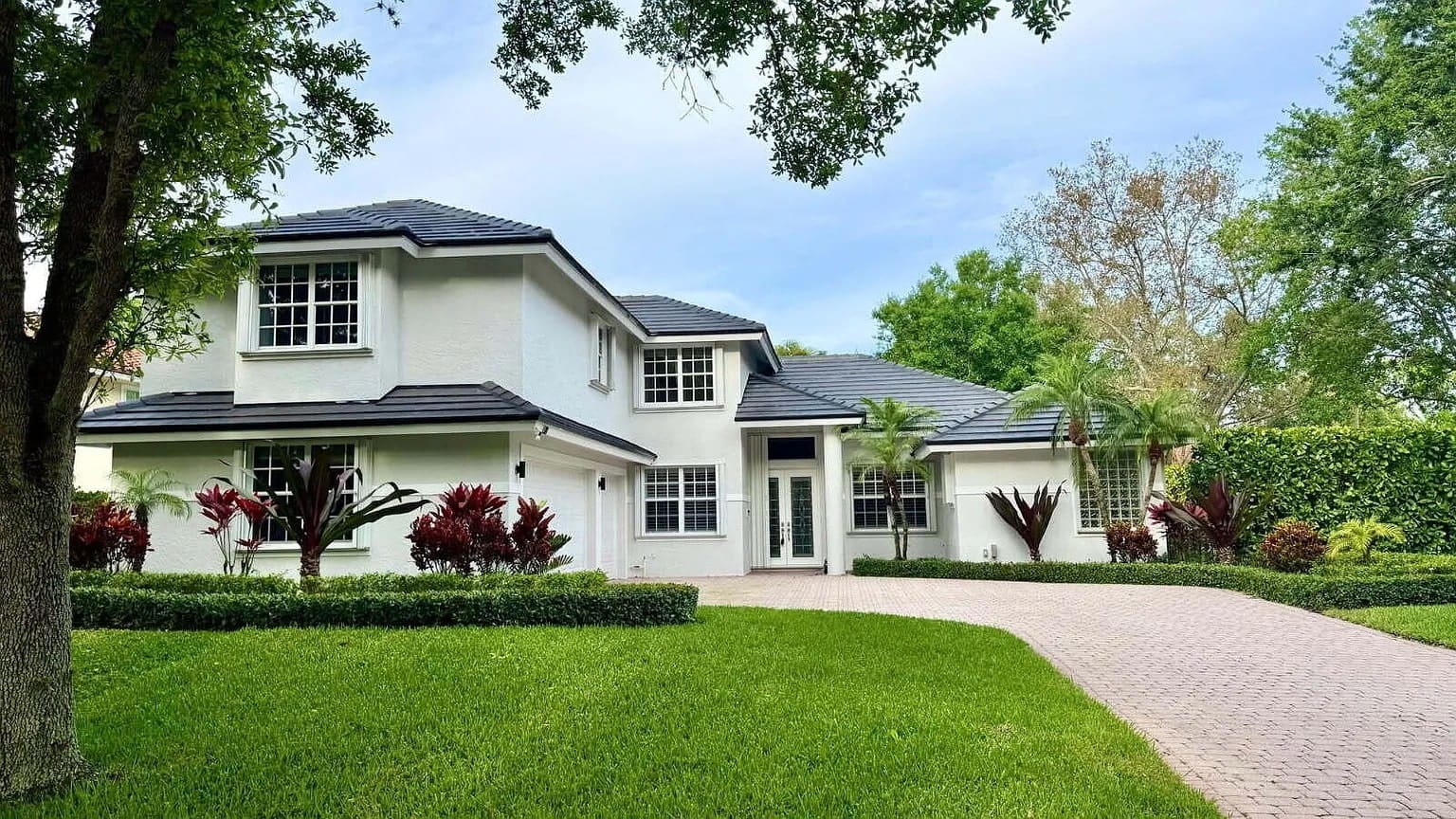 Two-story white house with a dark roof, surrounded by trees and a brick driveway.