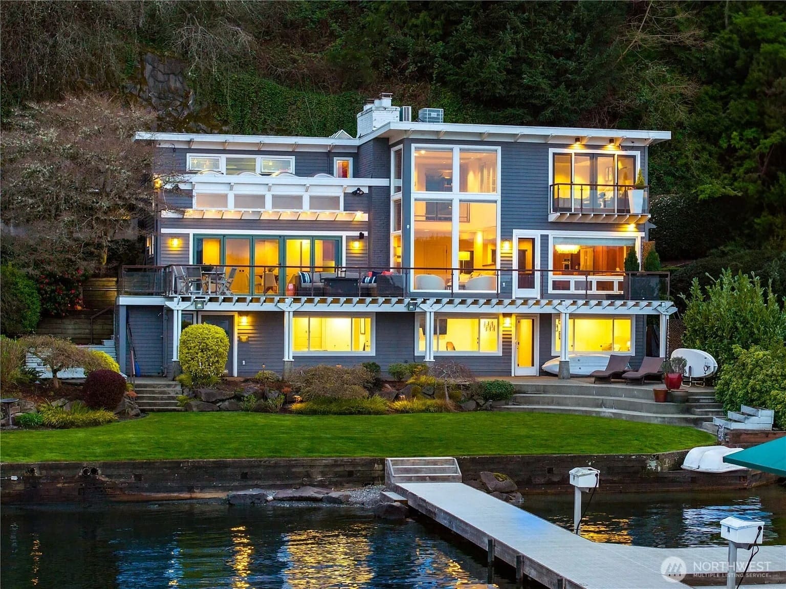Modern lakeside house with large windows, multiple balconies, and a dock extending into the water.