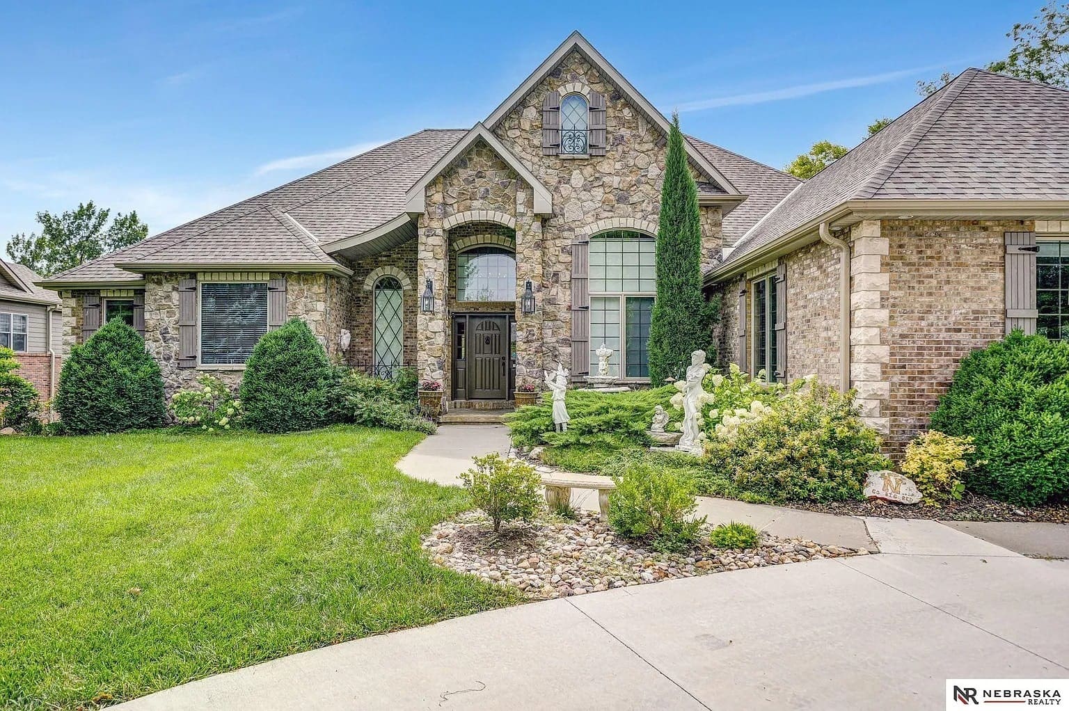 Stone and brick house with manicured lawn, shrubs, and decorative statues near the entrance.