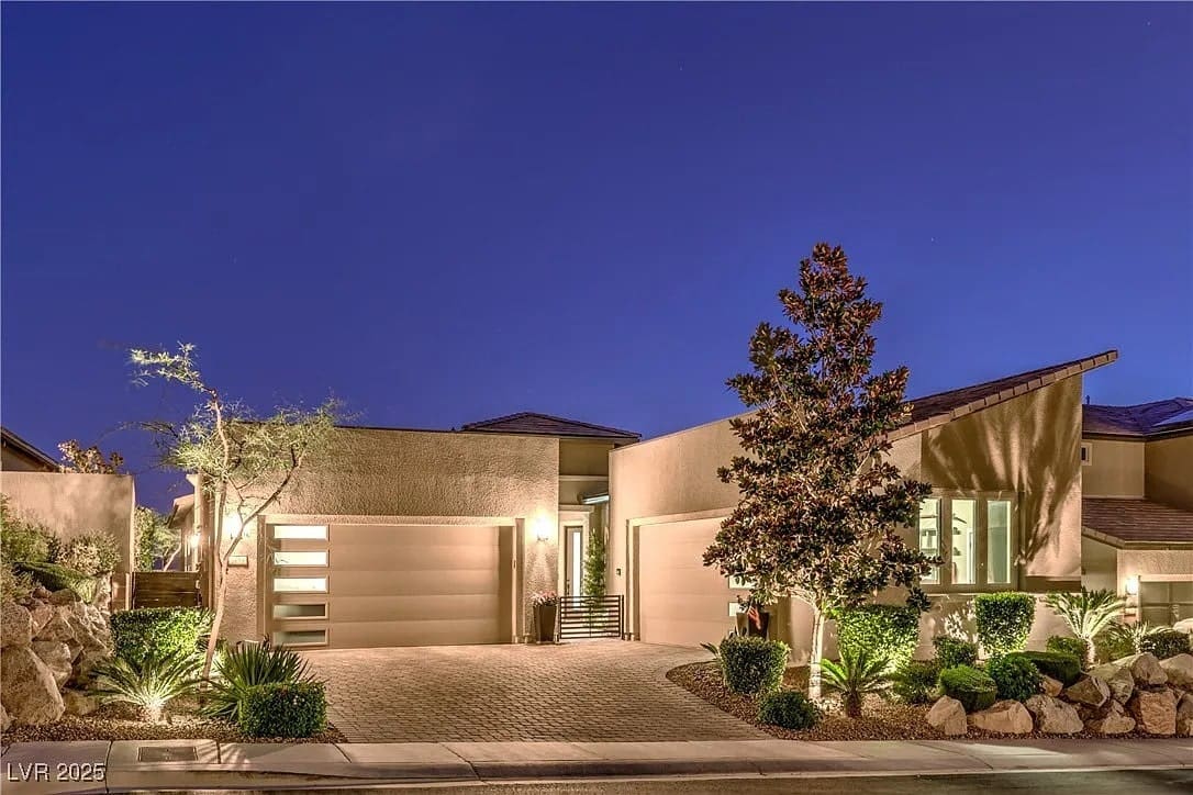 Modern house with large windows and garage, illuminated at night, surrounded by landscaped plants.