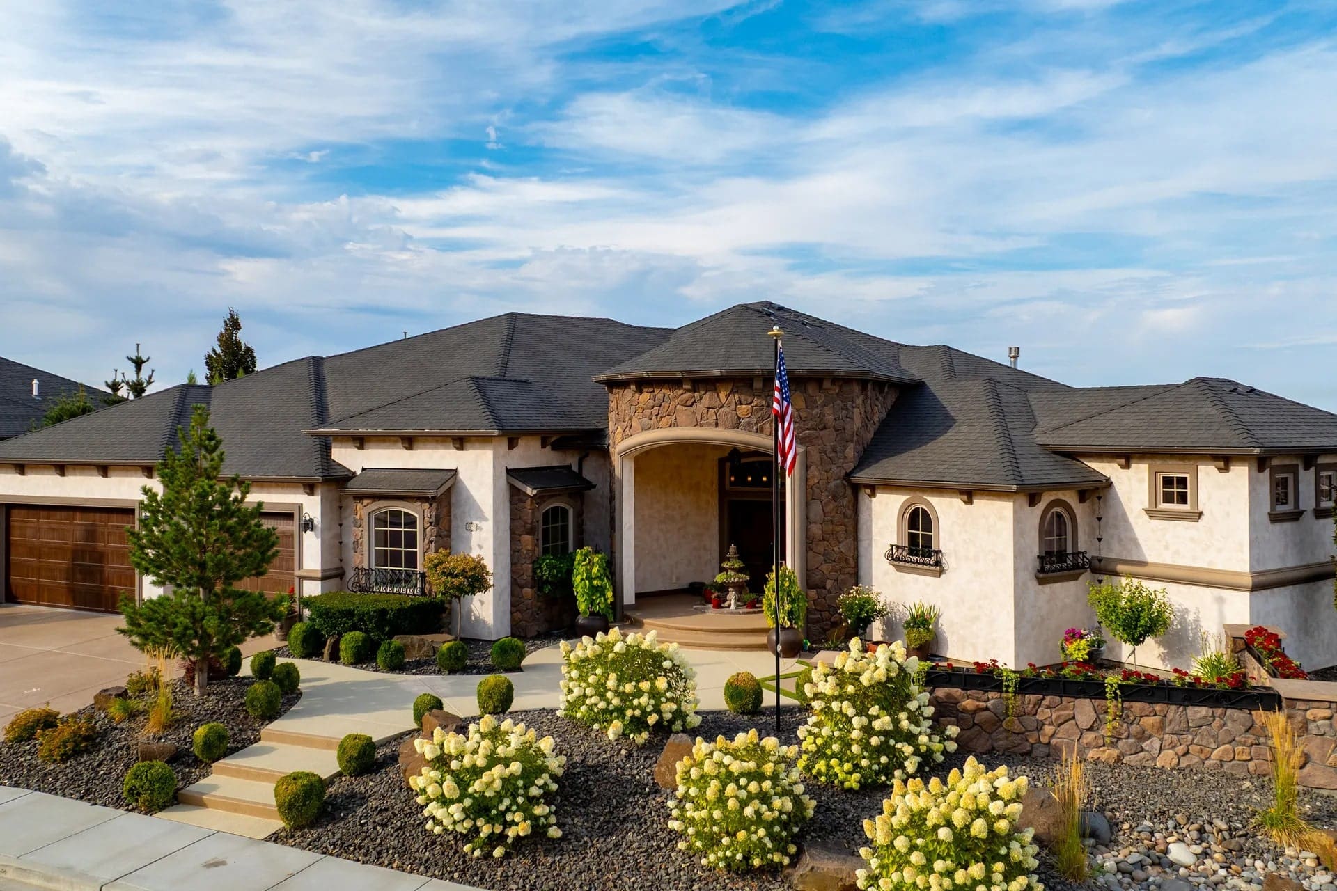 Large, elegant house with stone accents, manicured landscaping, and an American flag in front.