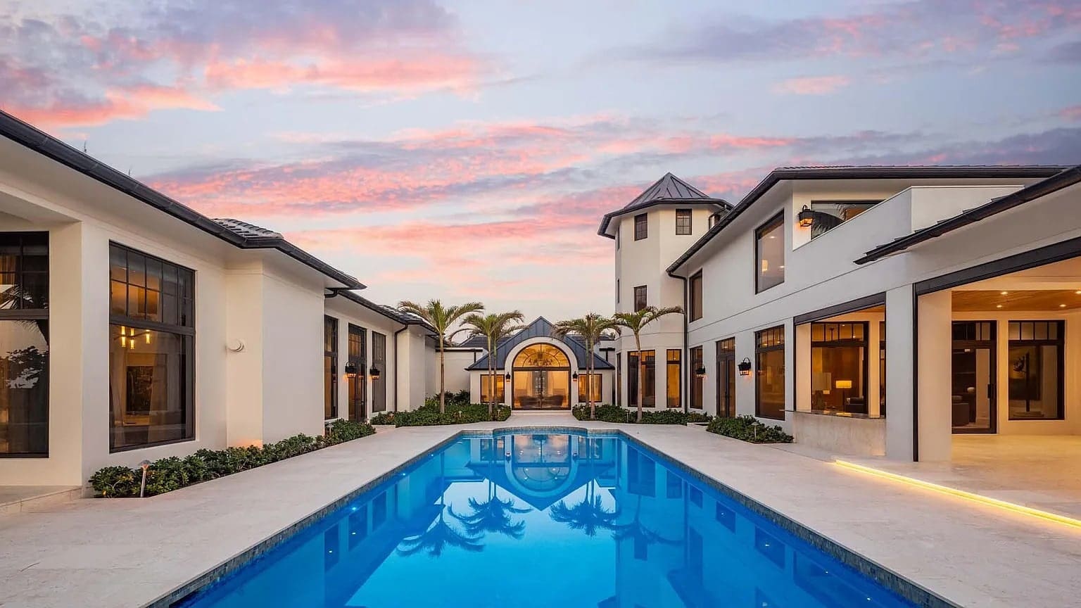 Luxury mansion with a central pool, surrounded by palm trees, under a colorful sunset sky.
