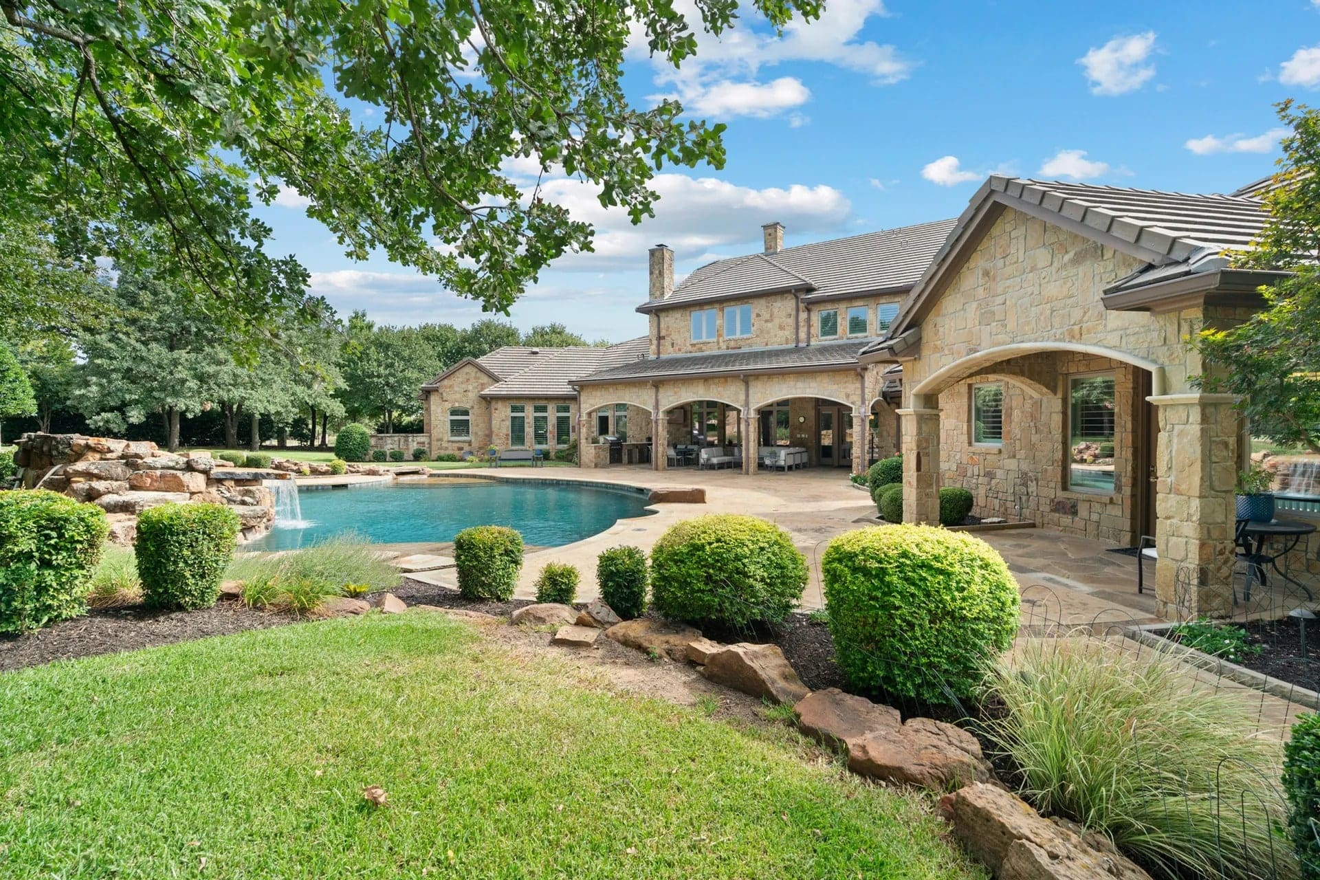Luxurious stone house with a large backyard pool, surrounded by greenery and trees.