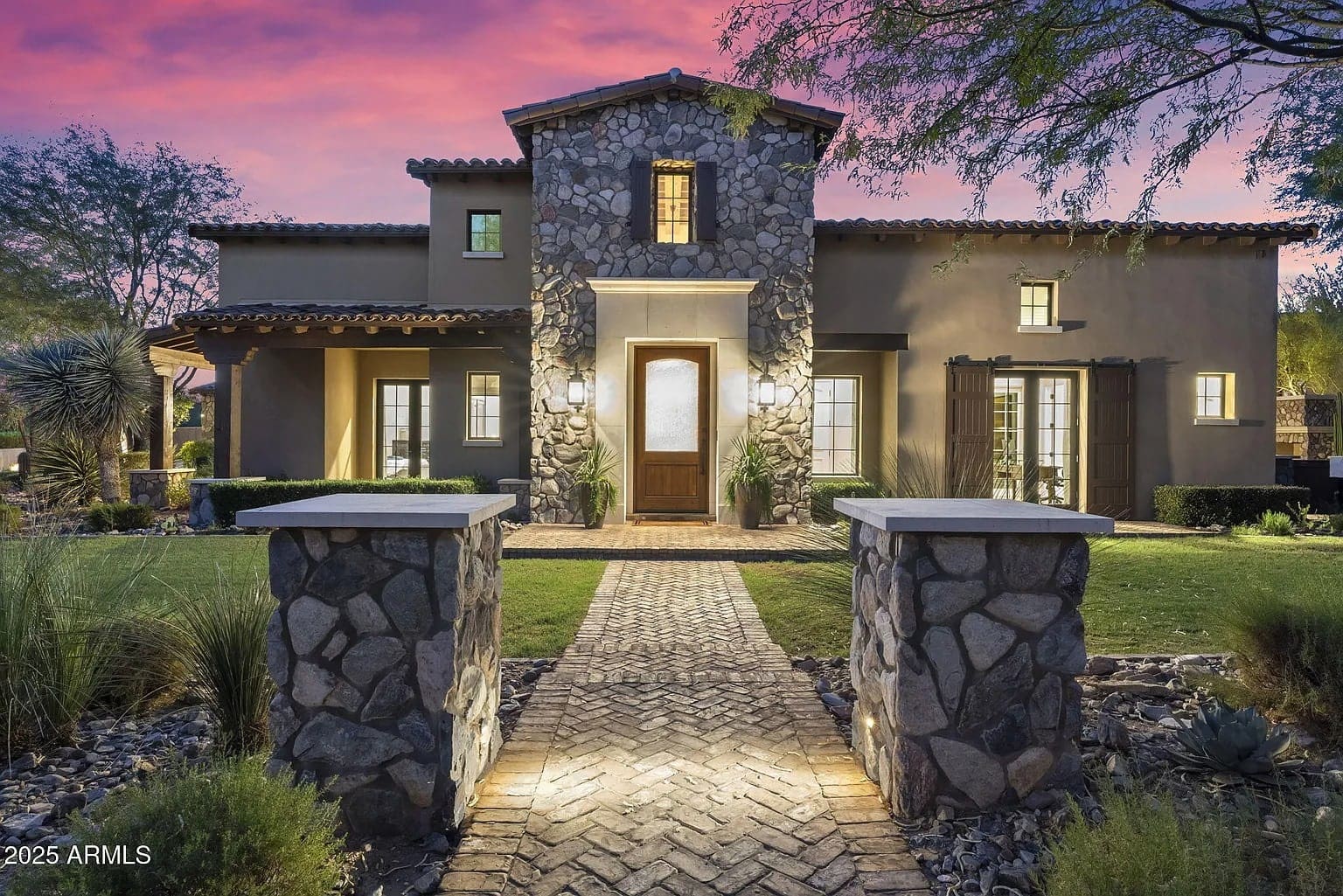 Stone and stucco house with a central tower, lit pathway, and landscaped yard at sunset.