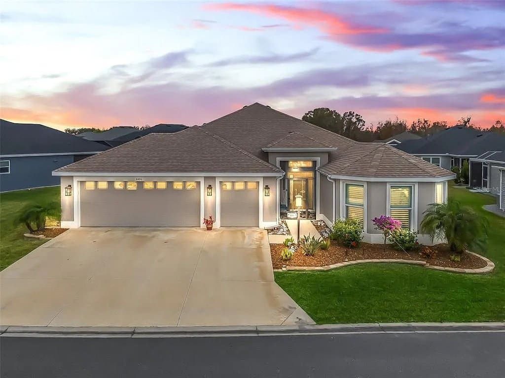 Modern single-story house with a three-car garage, landscaped yard, and sunset sky backdrop.
