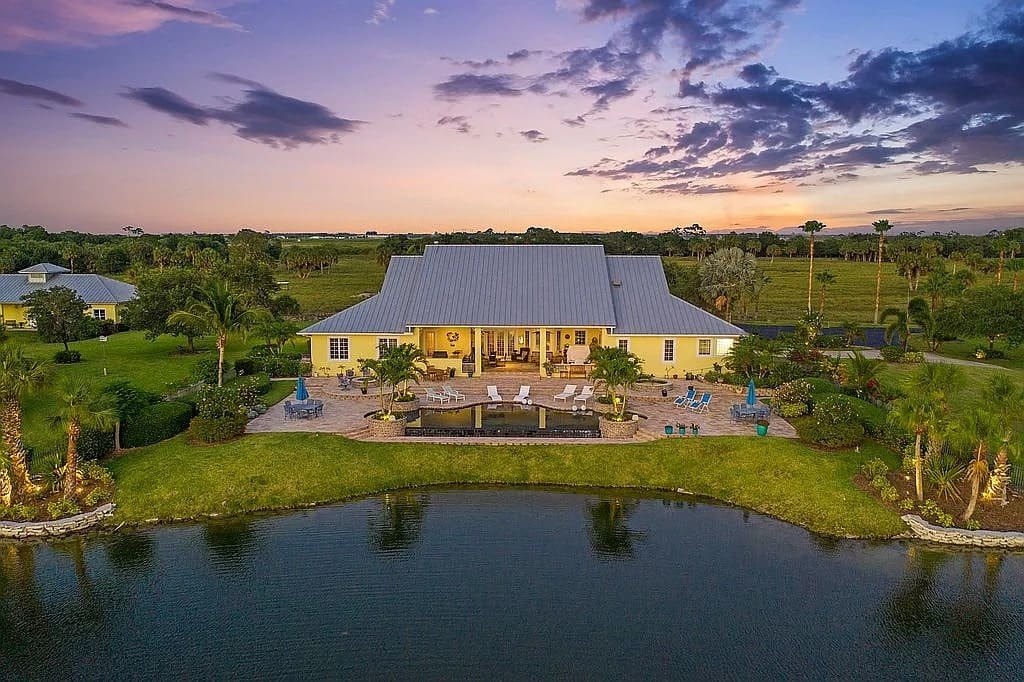 Large yellow house with a metal roof, patio, and pool, overlooking a pond at sunset.