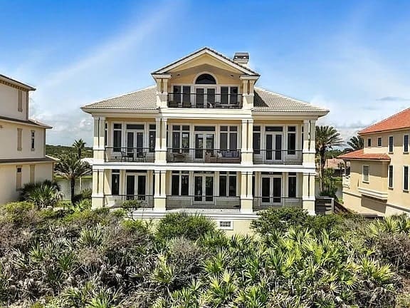 Large, three-story beachfront house with balconies, surrounded by greenery, under a clear blue sky.