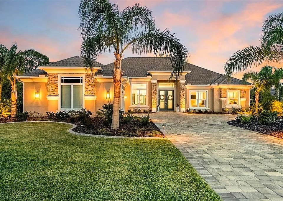 Single-story house with palm trees, illuminated at dusk, featuring a paved driveway and manicured lawn.