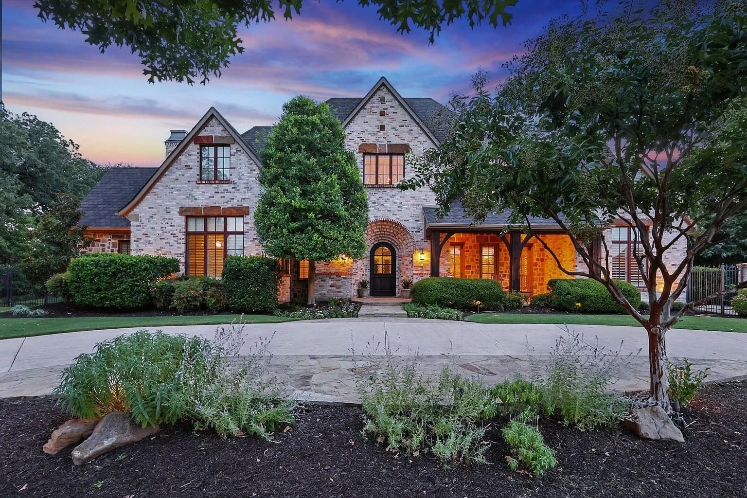 Large brick house with peaked roof, surrounded by trees and landscaped garden, at sunset.