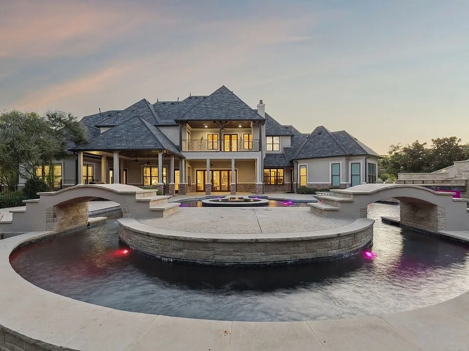 Luxury mansion with large pool and bridges, illuminated at dusk.