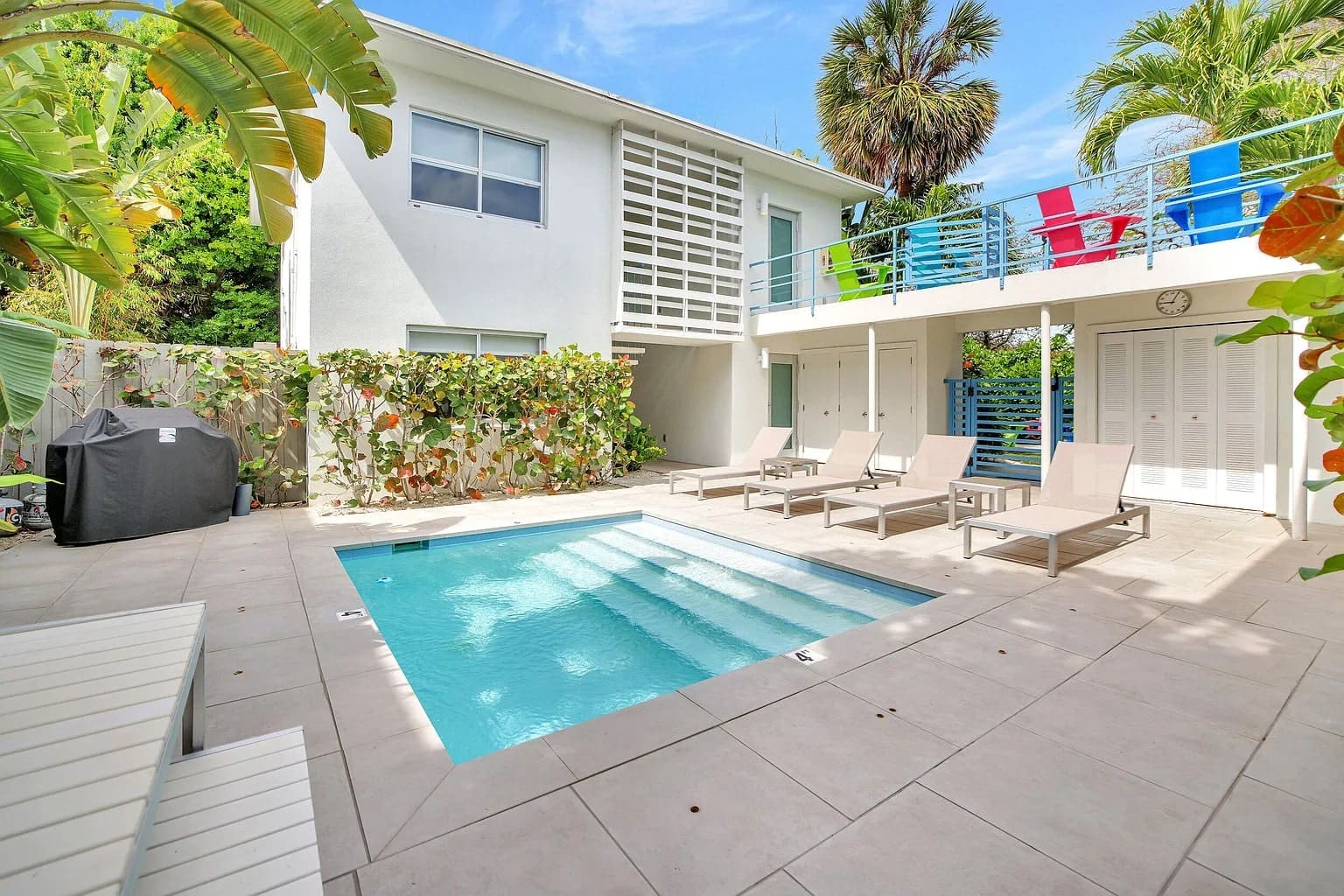 Modern backyard with a small pool, lounge chairs, and tropical plants surrounding a white two-story house.
