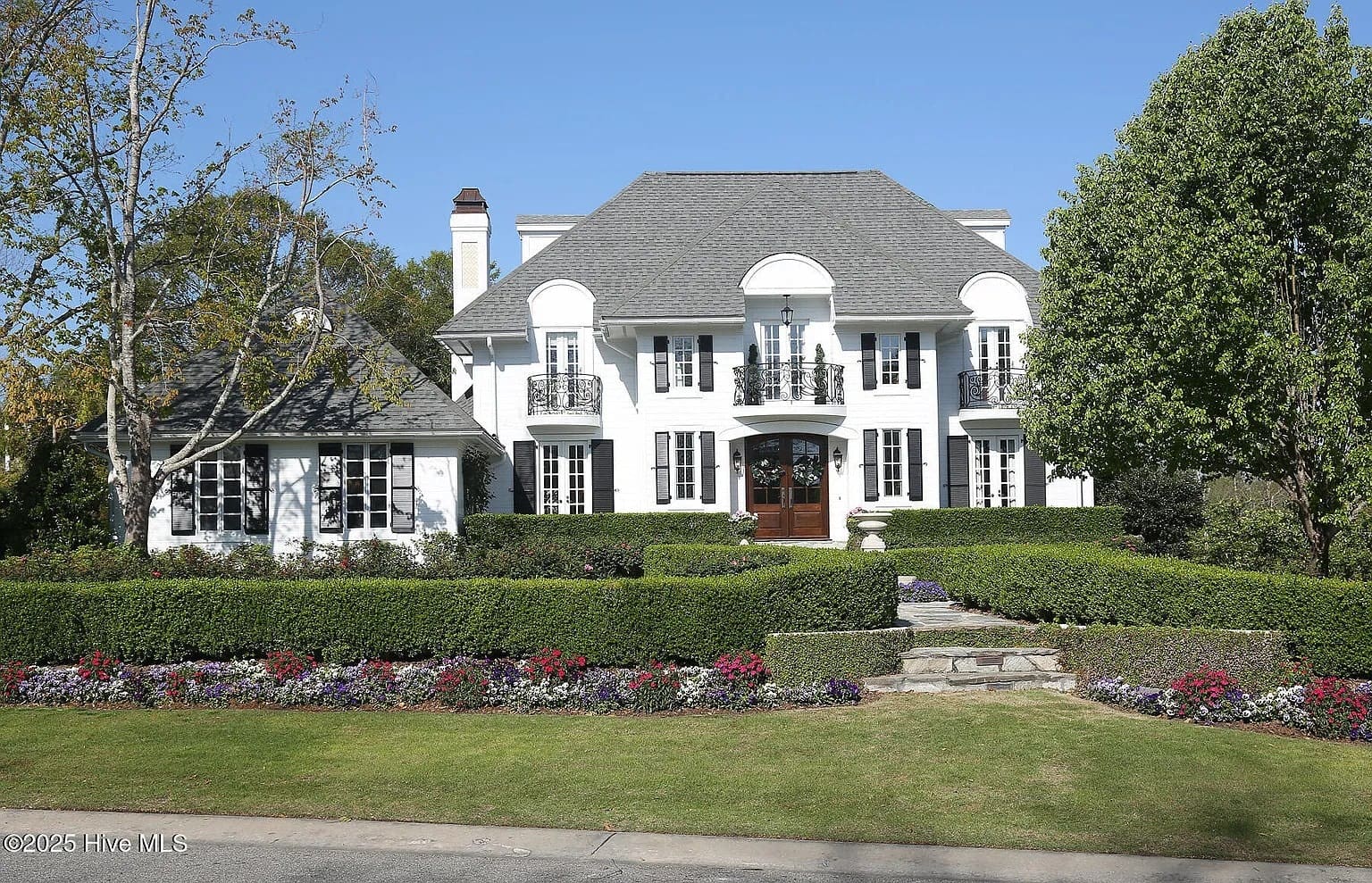 Large white two-story house with manicured hedges, colorful flowers, and a stone pathway.