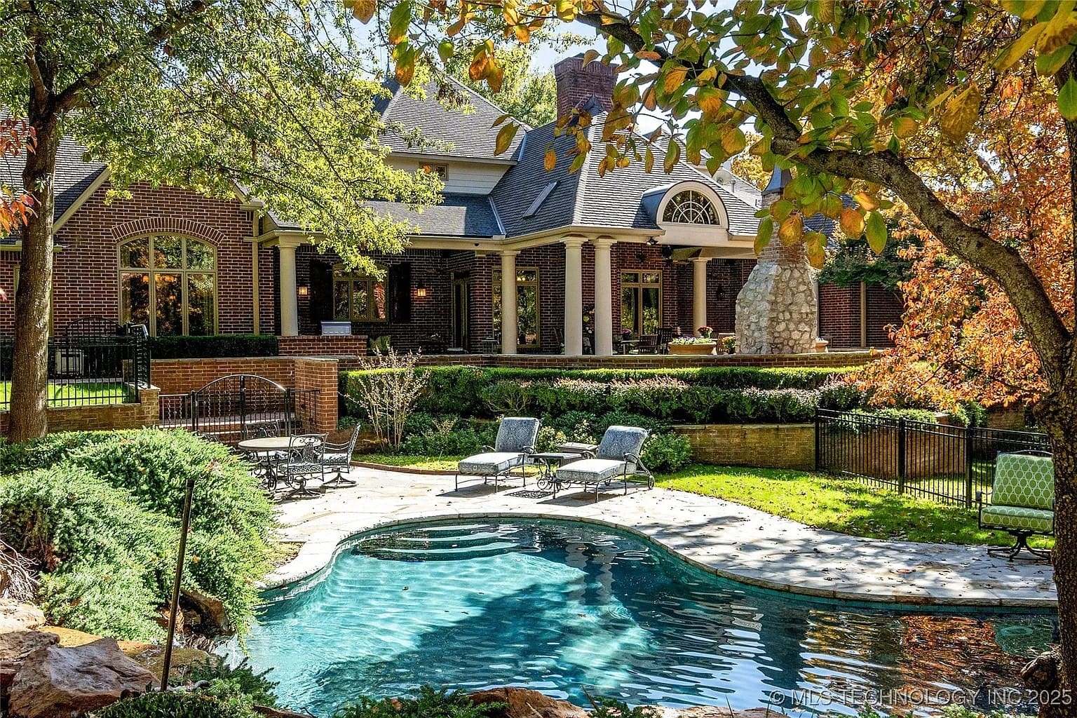 Backyard view of a brick house with a pool, lounge chairs, and autumn trees.