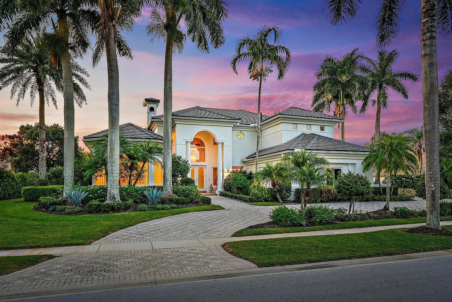 Large, elegant house with palm trees, lit warmly at sunset, featuring a curved driveway and manicured lawn.