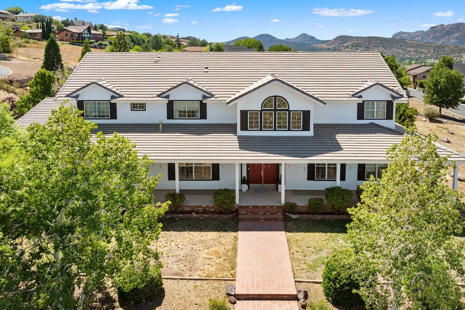 Large two-story house with a gray roof, surrounded by trees, set in a hilly landscape.
