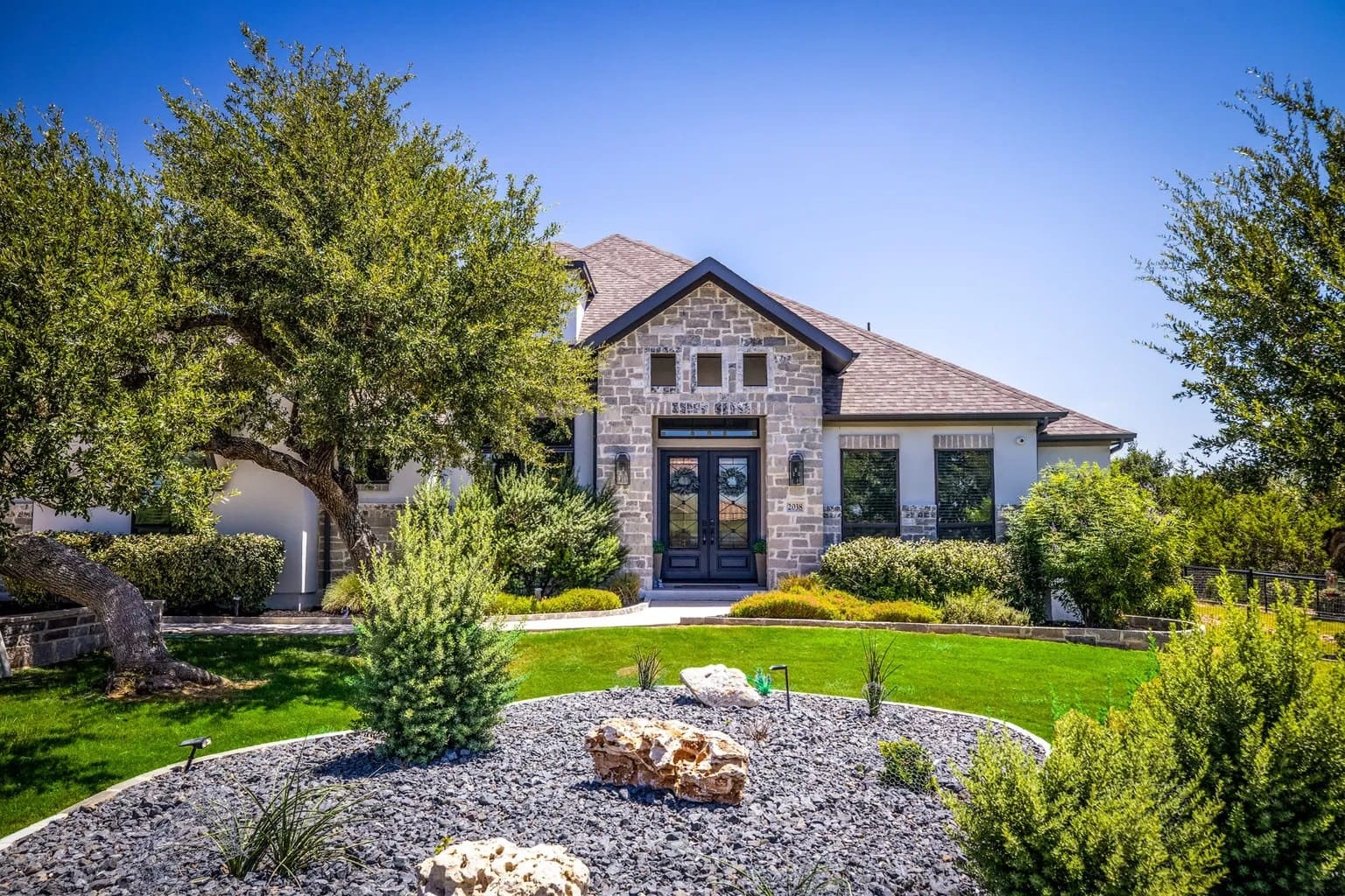 Modern stone house with large windows, surrounded by landscaped garden and trees under a clear blue sky.
