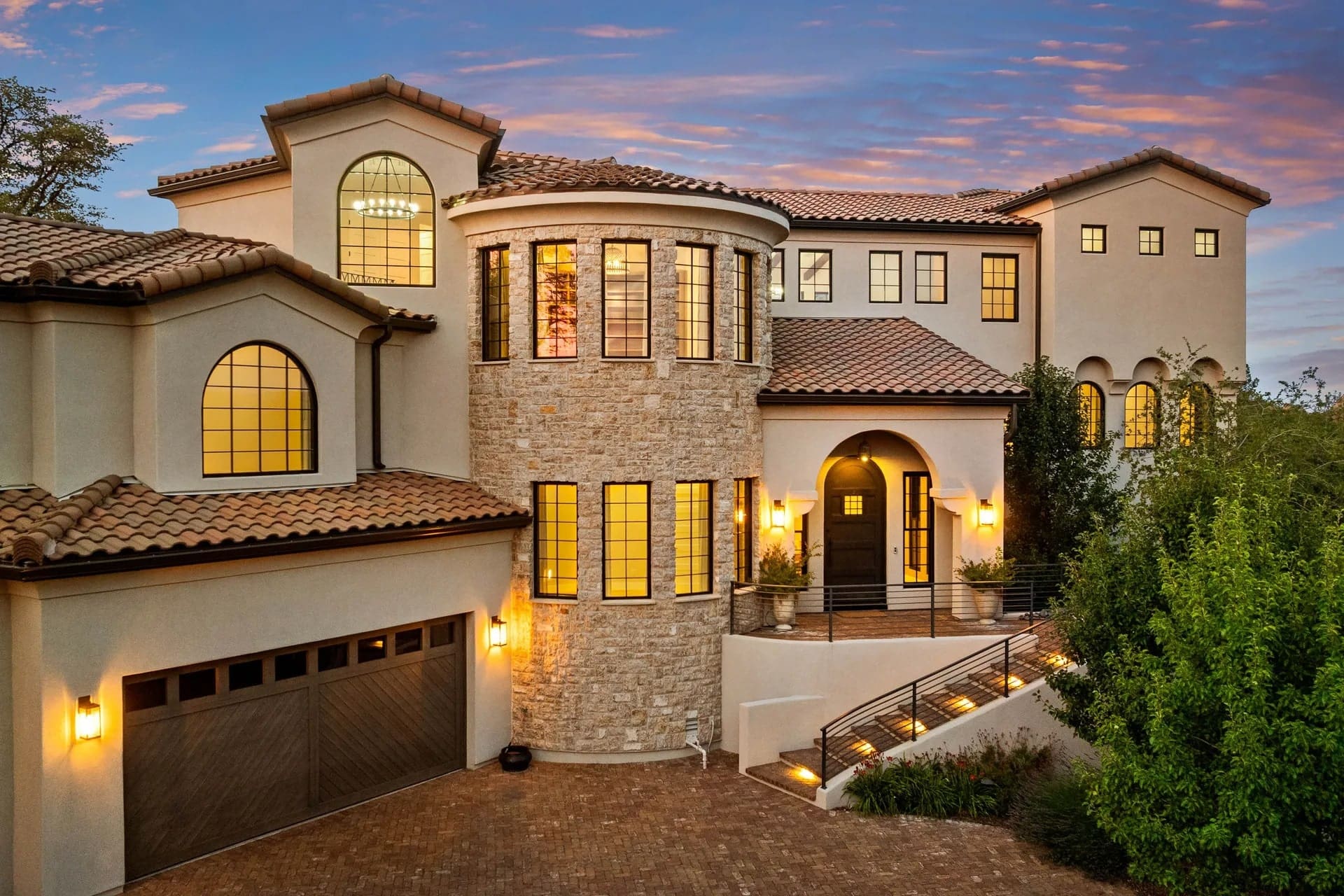 Large Mediterranean-style house with stone facade, arched windows, and warm exterior lighting at sunset.