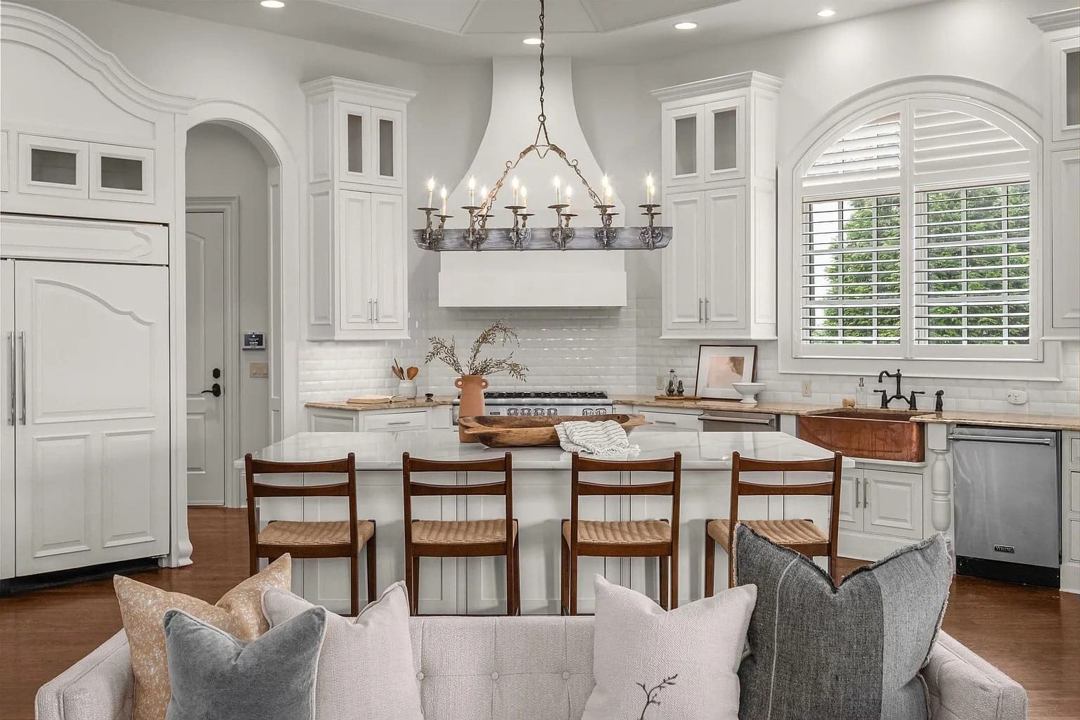 Spacious white kitchen with island, wooden chairs, chandelier, and large windows with shutters.
