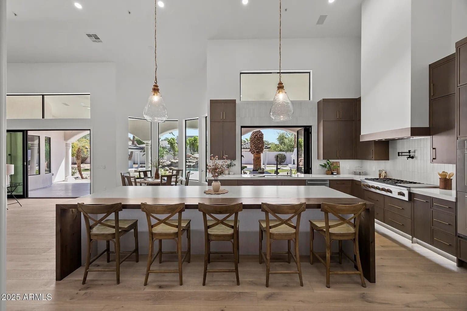 Modern kitchen with large island, wooden chairs, pendant lights, and view of outdoor patio.
