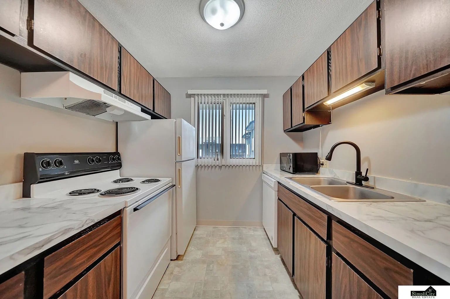 Narrow kitchen with dark wood cabinets, white appliances, and a window at the end.
