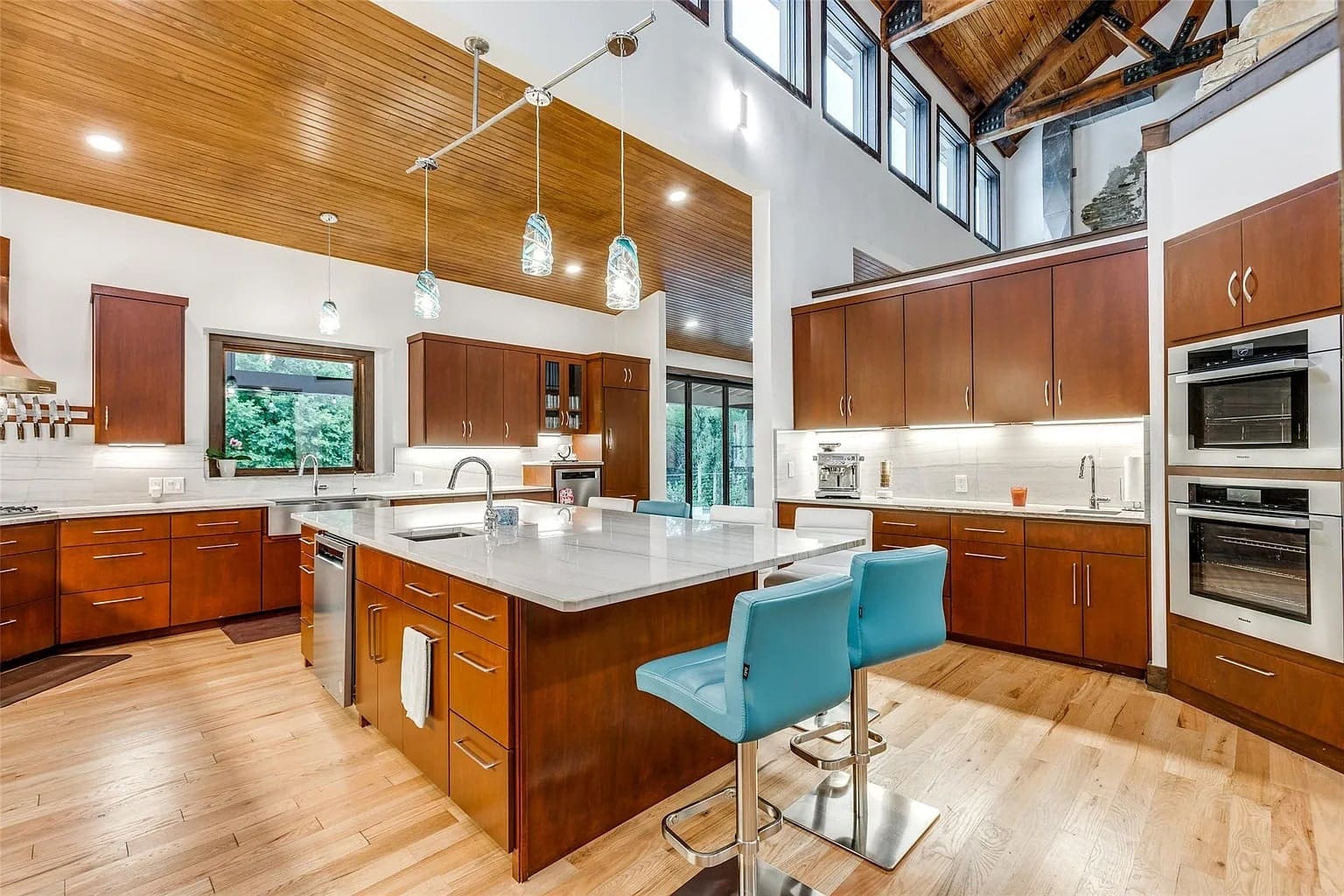 Spacious modern kitchen with wooden cabinets, large island, blue barstools, and stainless steel appliances.
