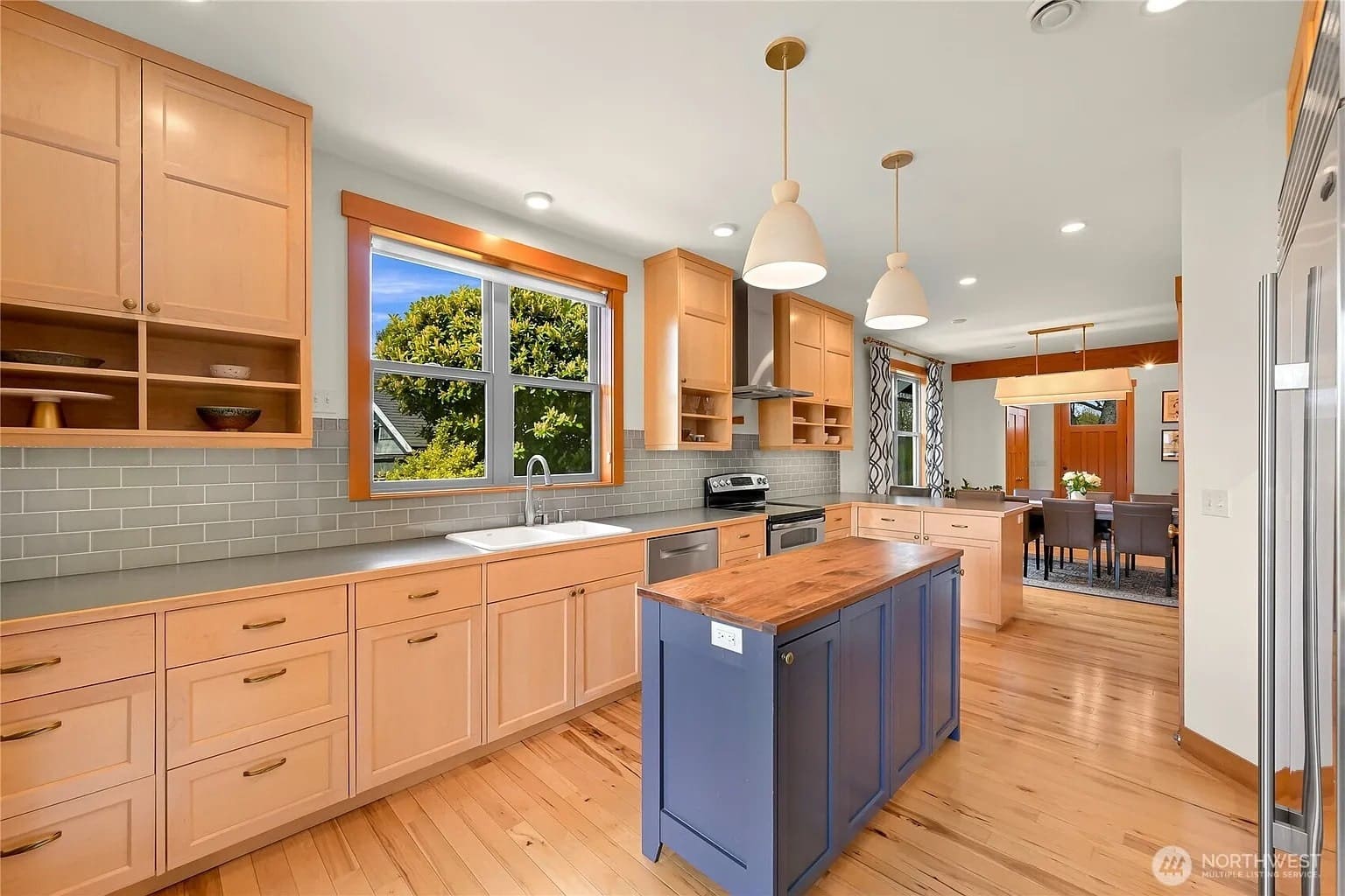 Modern kitchen with wooden cabinets, blue island, pendant lights, and dining area in the background.

