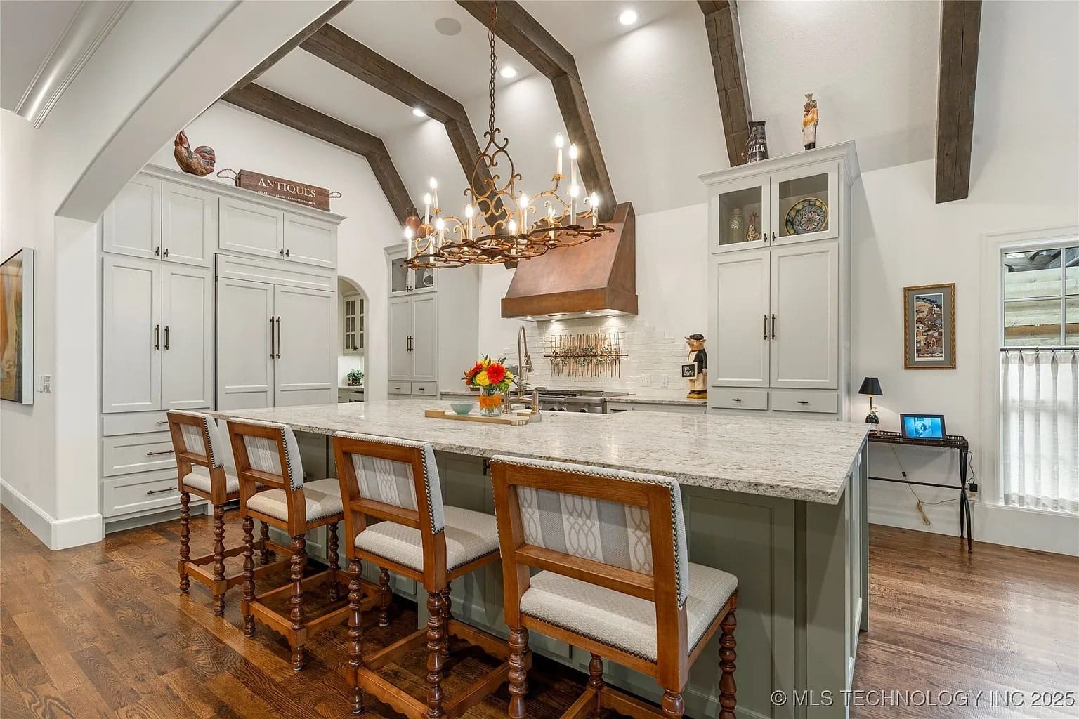 Spacious kitchen with wooden beams, large island, chandelier, and bar stools.
