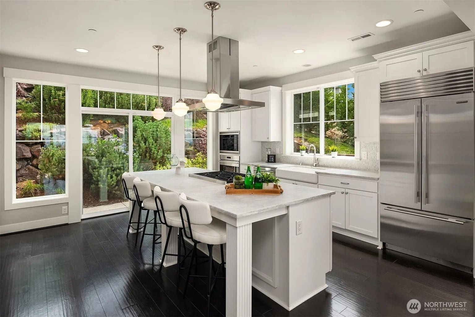 Modern kitchen with white cabinets, large island, stainless steel appliances, and large windows overlooking greenery.
