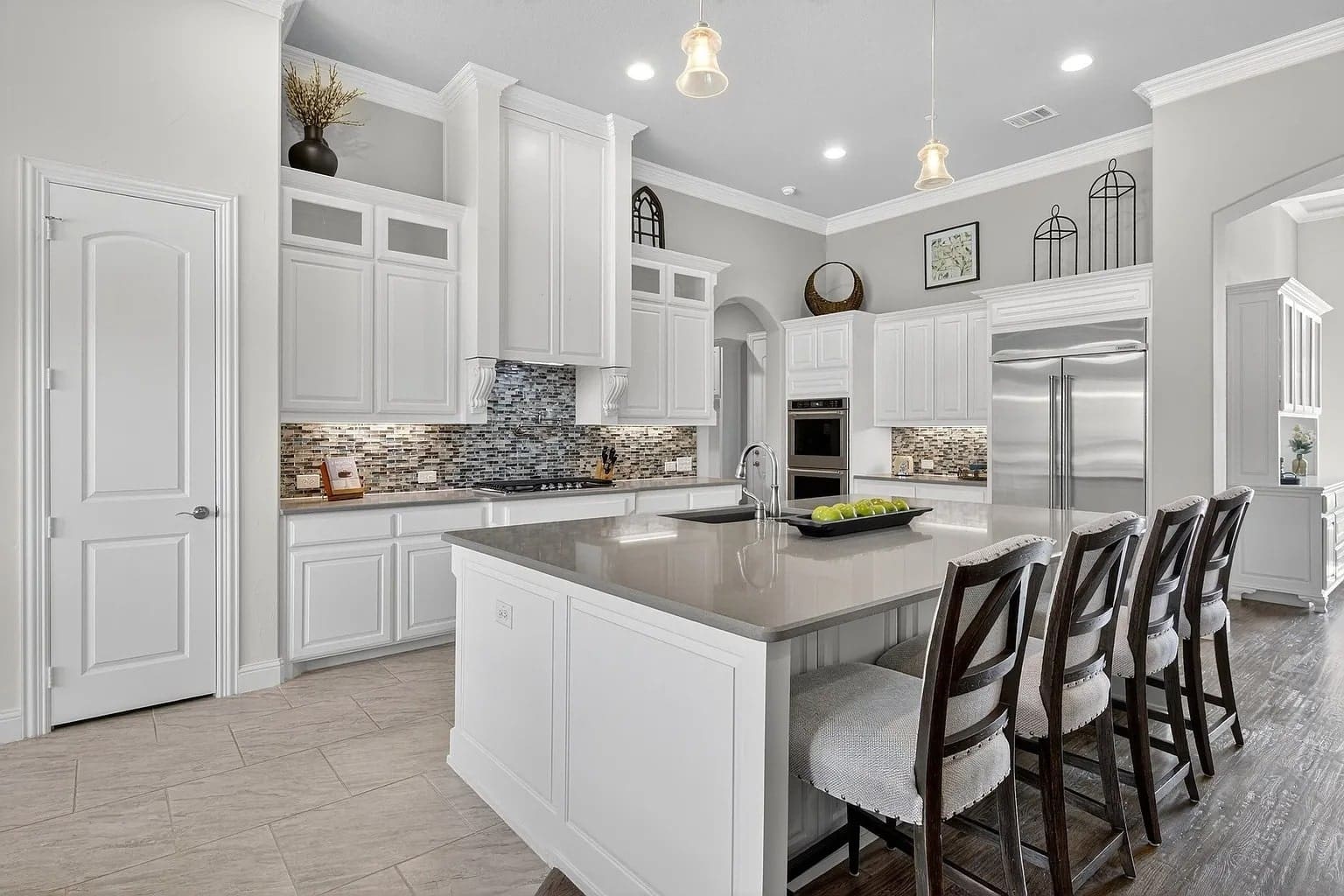 Modern kitchen with white cabinets, large island, bar stools, stainless steel appliances, and tile backsplash.
