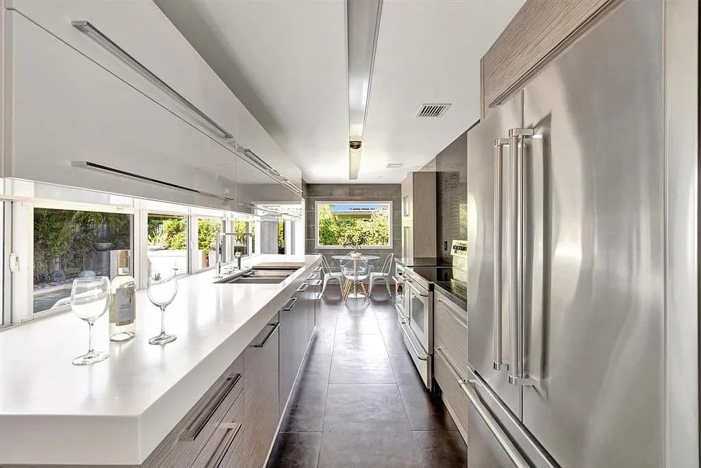 Modern kitchen with stainless steel appliances, white countertops, and large windows overlooking a dining area.
