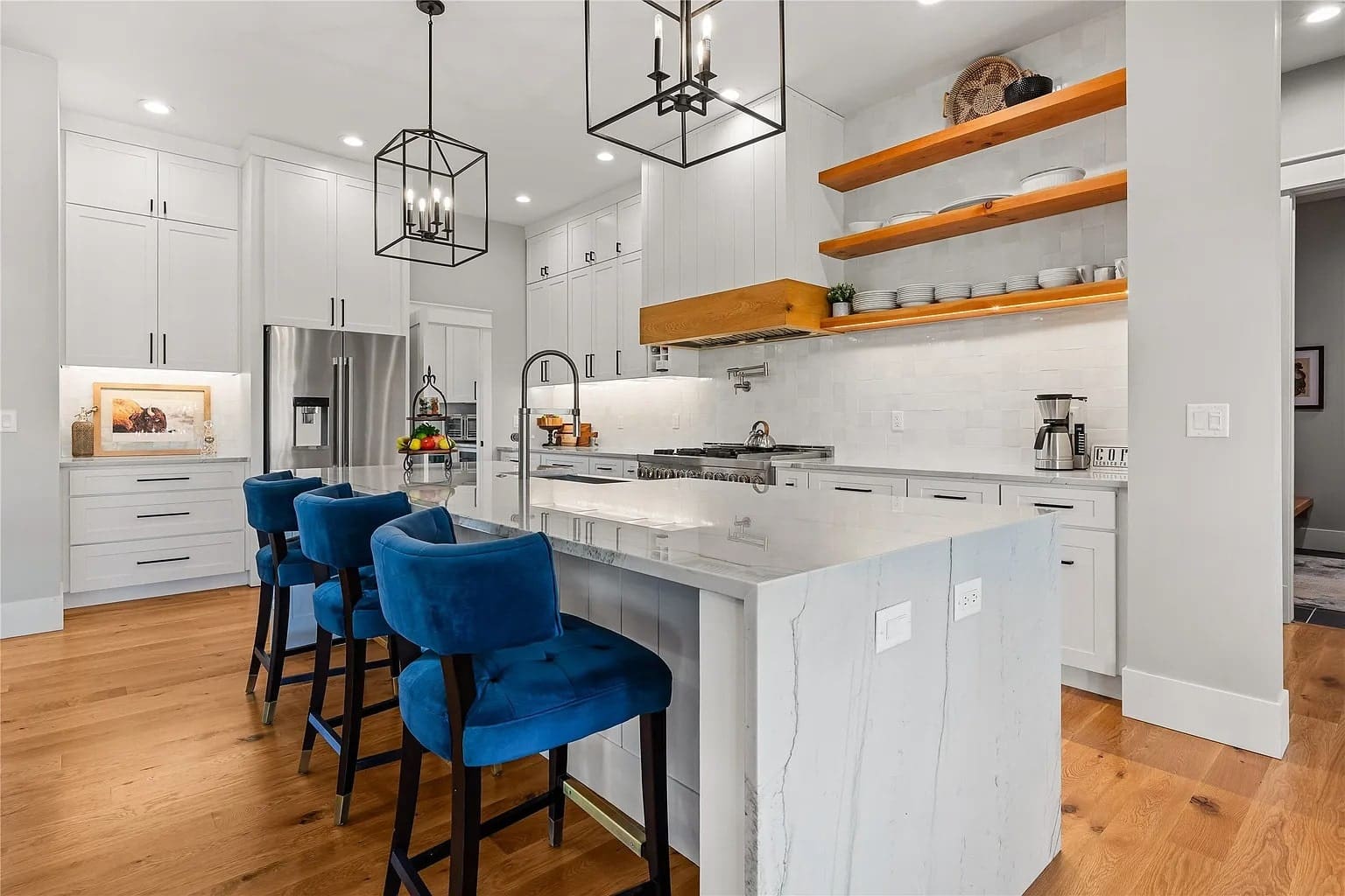 Modern kitchen with white cabinets, blue barstools, marble island, and wooden shelves.