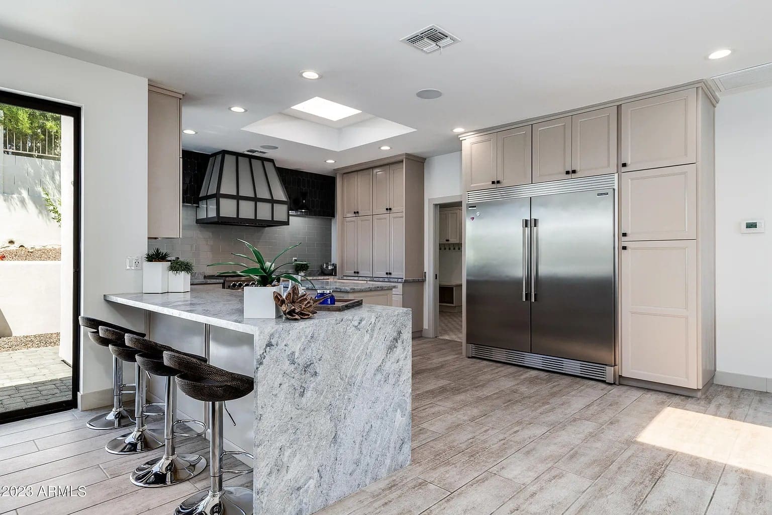 Modern kitchen with marble island, bar stools, large stainless steel fridge, and light wood cabinetry.