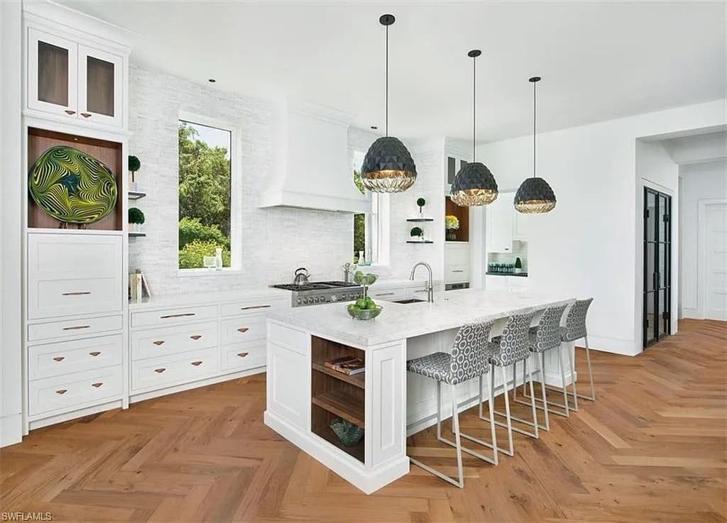 Modern kitchen with white cabinets, marble island, patterned stools, and pendant lights on herringbone wood floor.
