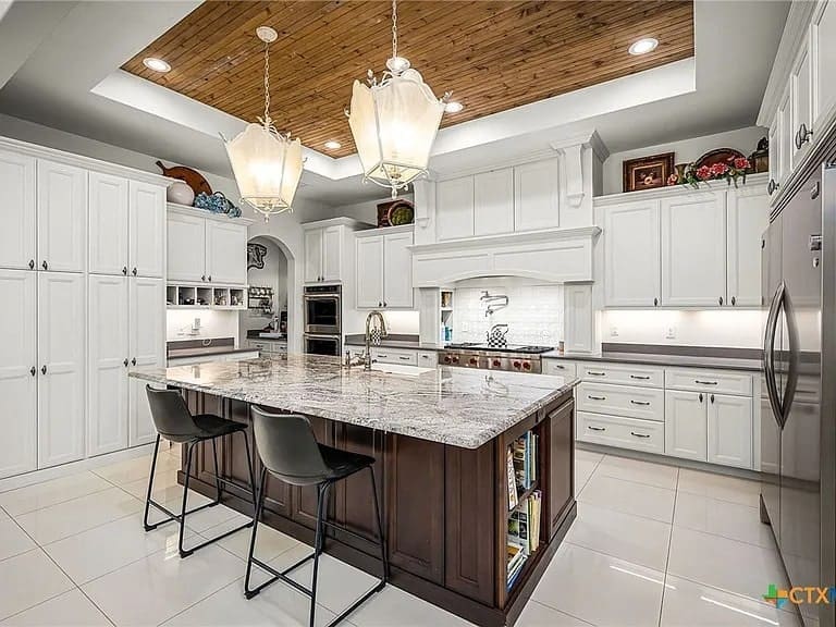 Modern kitchen with large marble island, dark wood base, white cabinets, and hanging lights.
