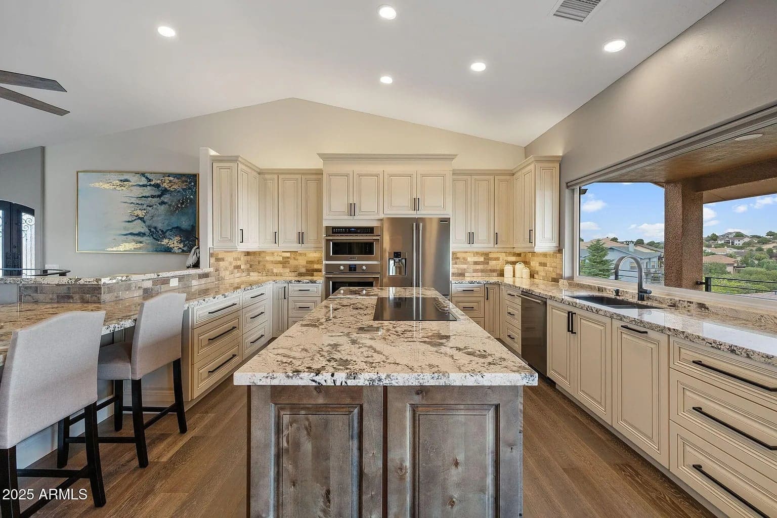 Modern kitchen with marble countertops, island, bar stools, stainless steel appliances, and large window view.

