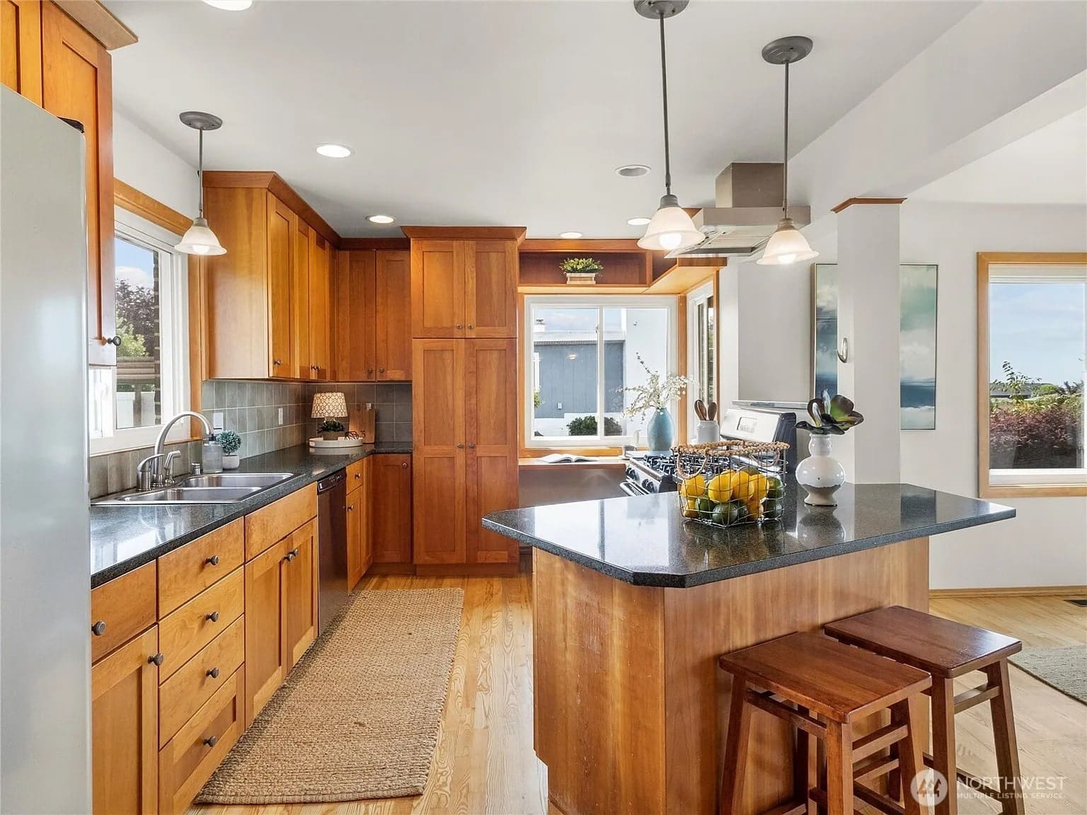 Modern kitchen with wooden cabinets, island with stools, stainless steel appliances, and large windows.
