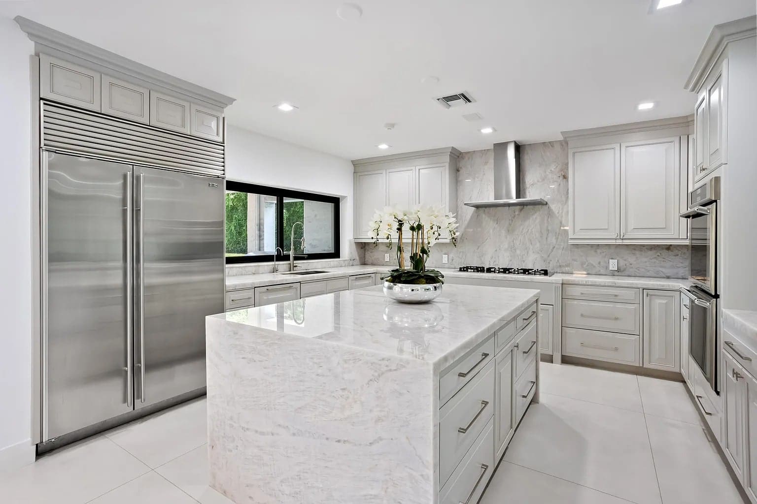 Modern kitchen with marble countertops, stainless steel appliances, and white cabinetry. Central island with orchids.