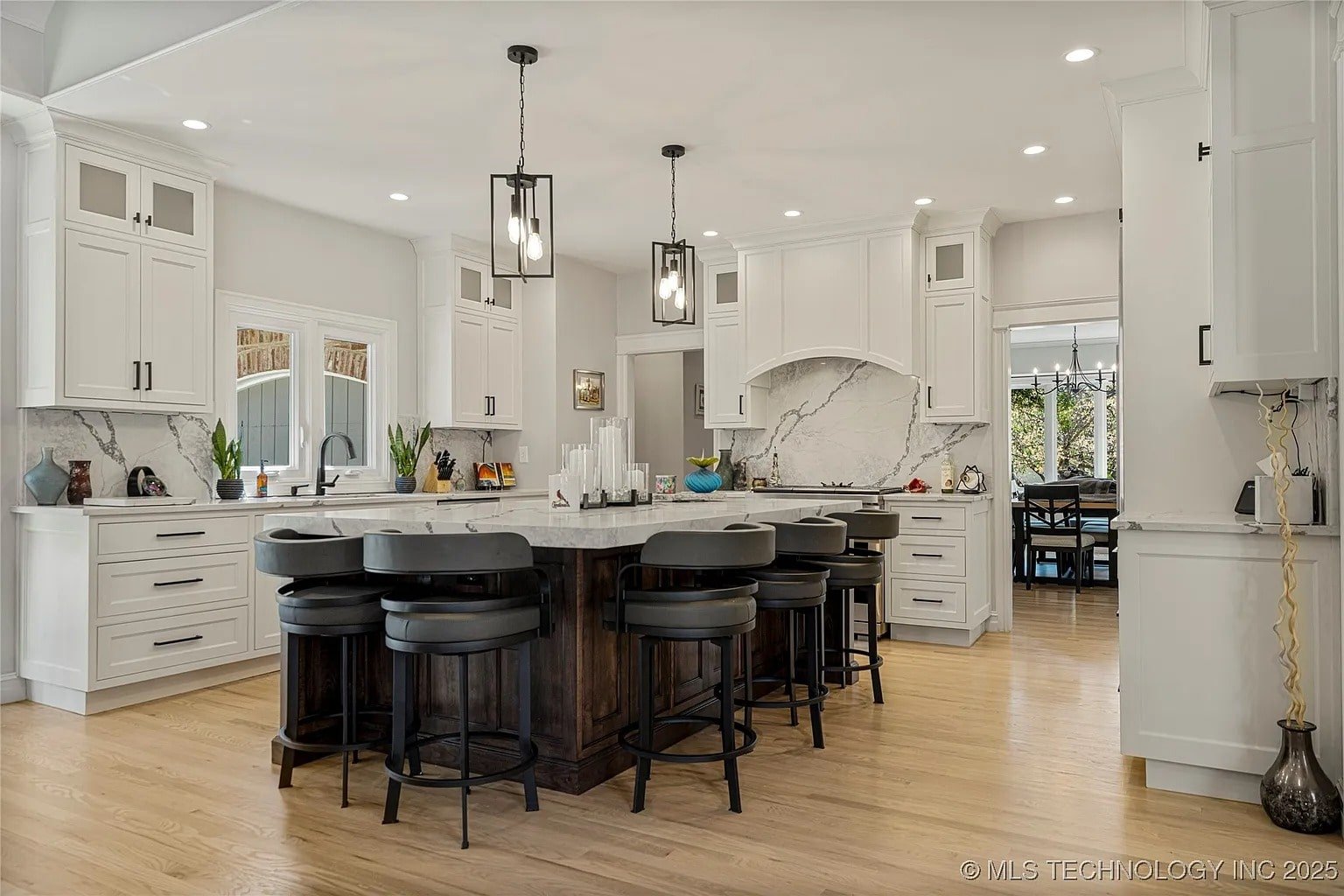 Modern kitchen with white cabinets, large island, black chairs, pendant lights, and hardwood floors.